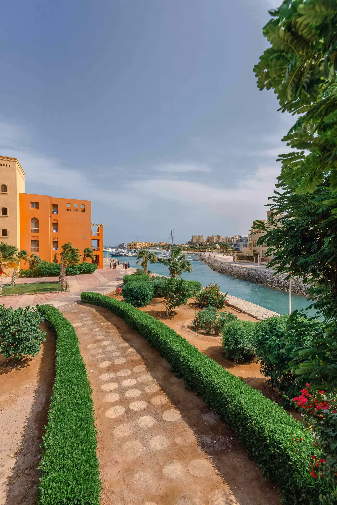 Hurghada marina waterway with manicured hedges, orange buildings, moored yachts, and palm trees under blue sky