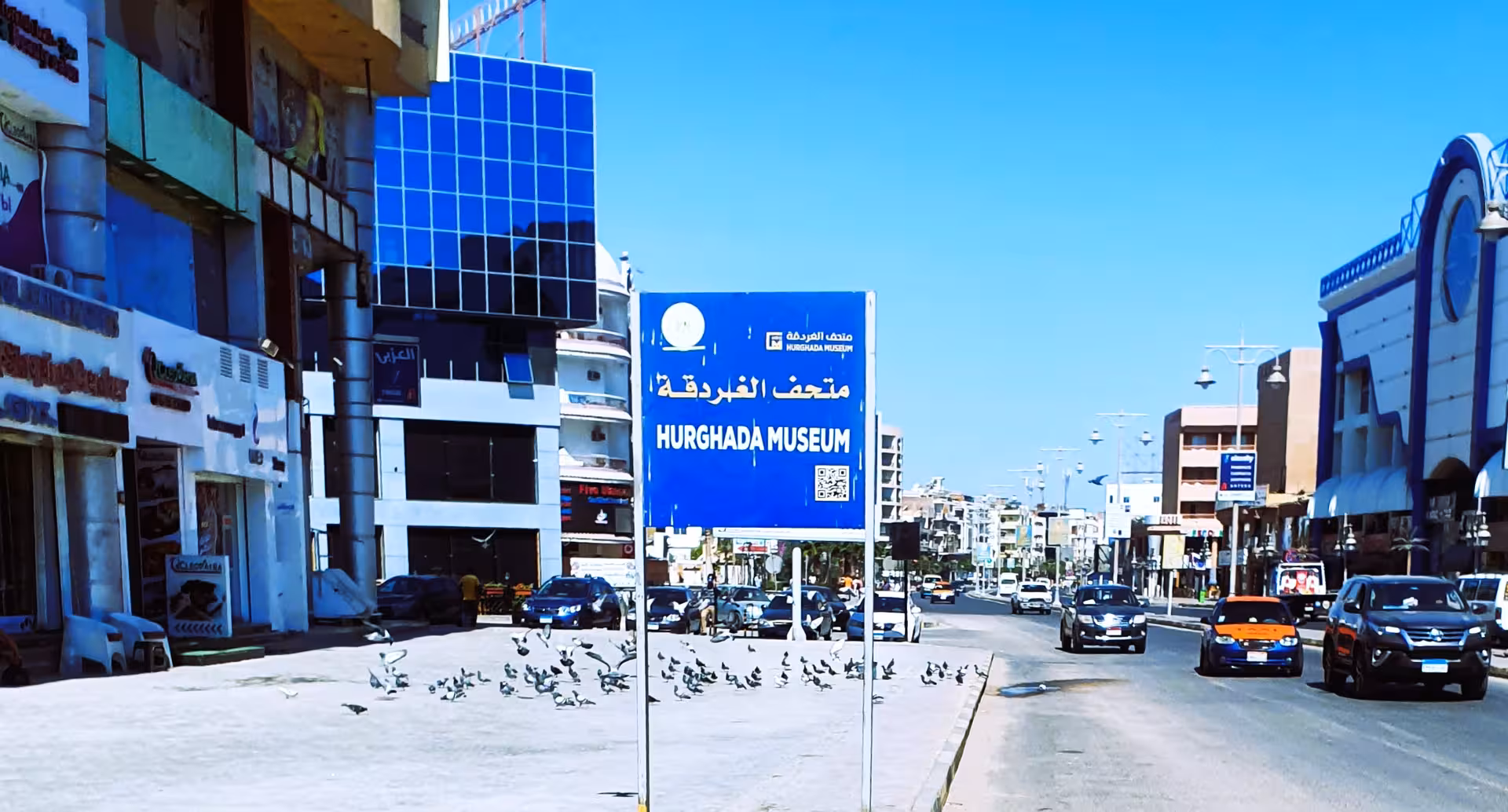 Hurghada Museum street sign in Arabic and English on a busy city road with shops and traffic