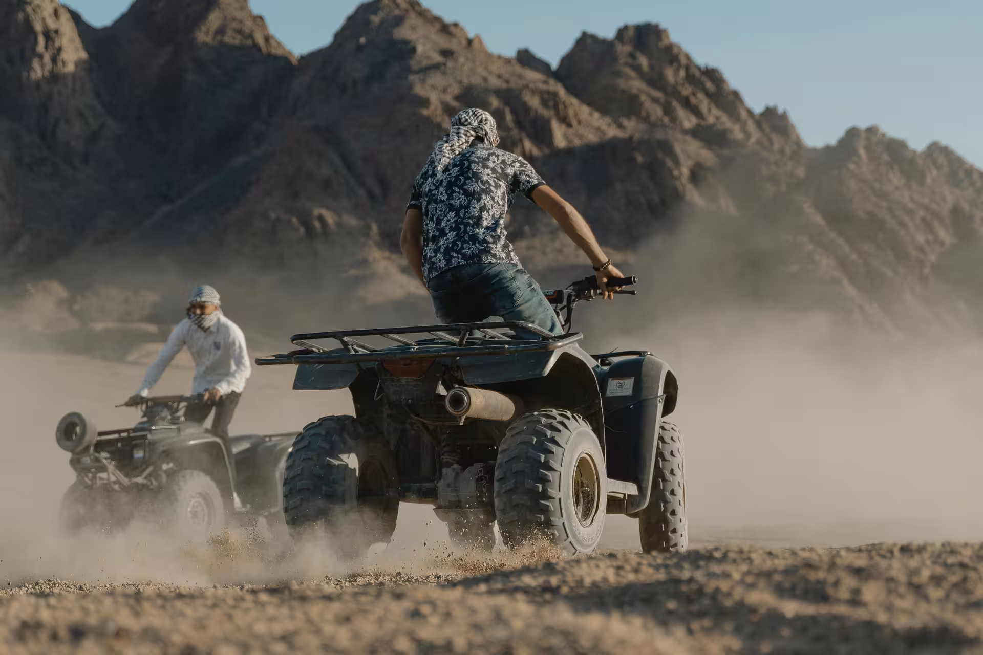 Two riders on quad bikes speeding through dusty desert terrain with rocky mountains in background, Hurghada
