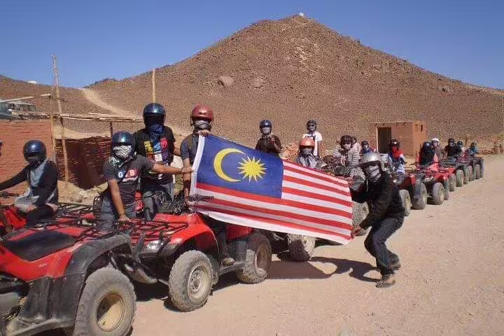 Quad bike desert safari group lined up at camp, riders holding Malaysian flag before sunset dune adventure
