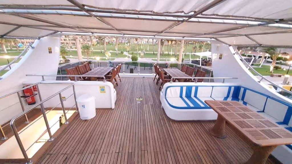 Upper deck of Hurghada tour boat with wooden flooring, white cushioned seating, dining tables, and resort view