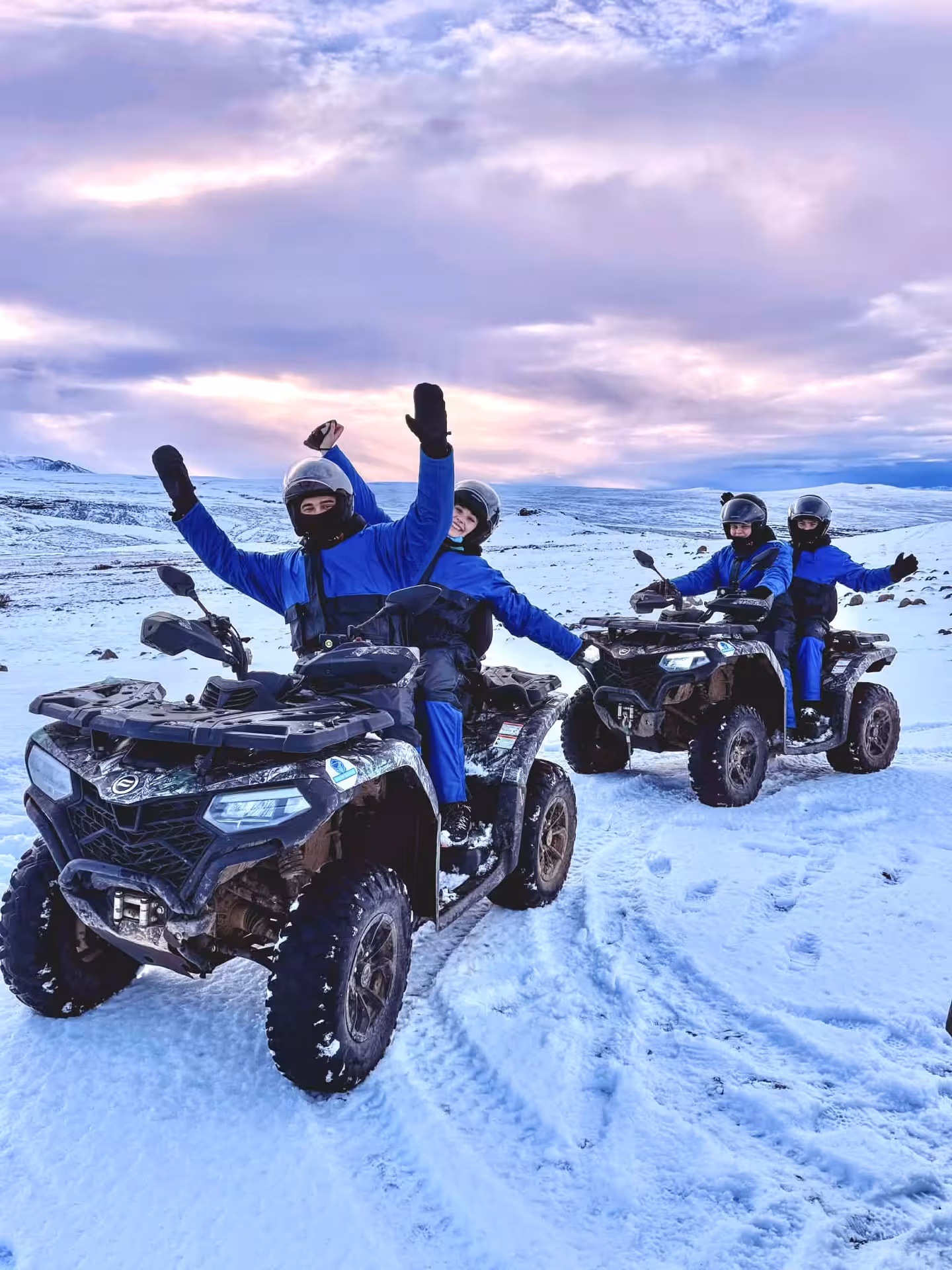 Four riders in blue suits on ATVs celebrating on snowy Husafell mountain landscape under pink sky