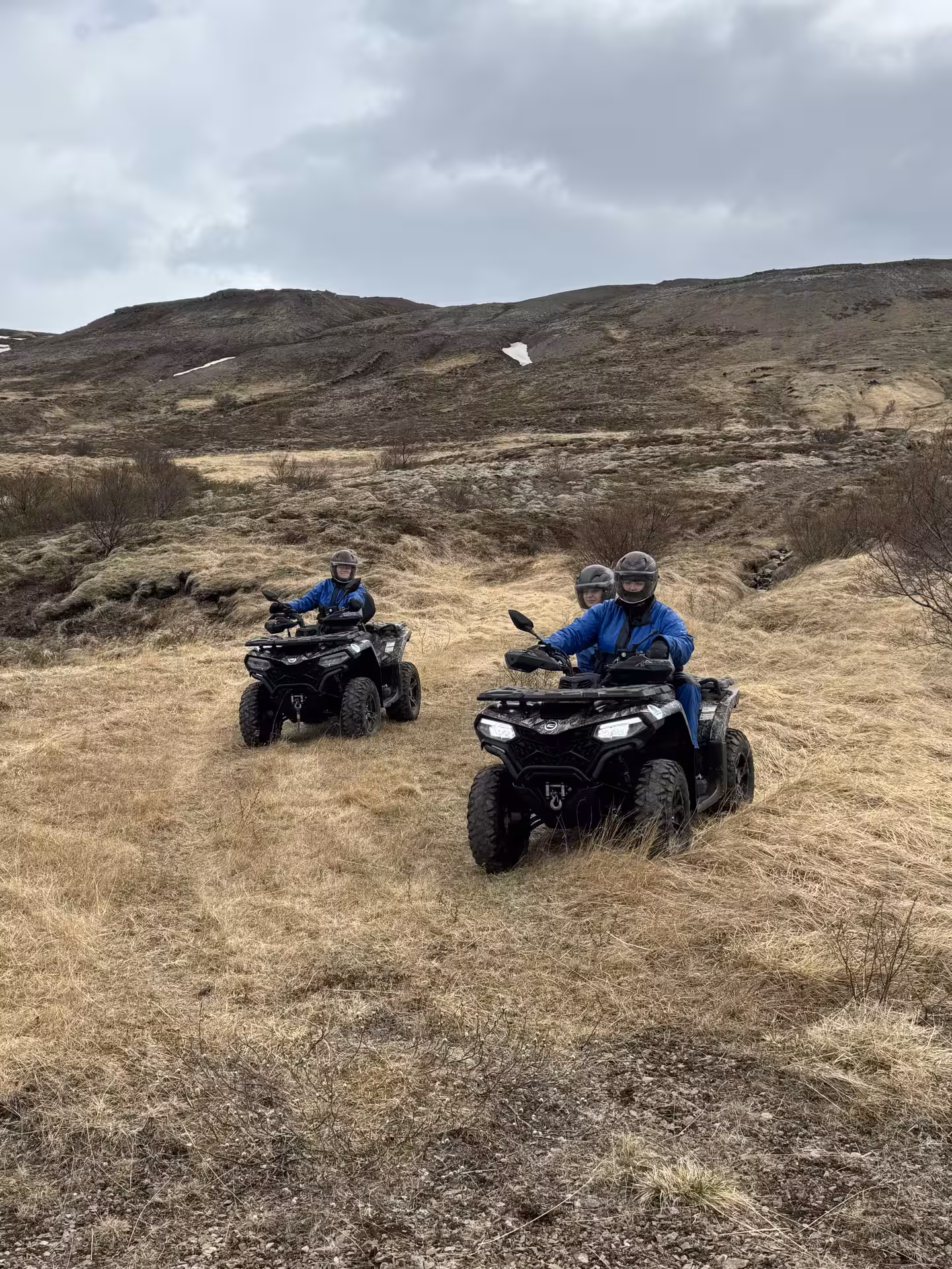 Two riders on black ATV quad bikes crossing grassy highland terrain with mountain views in Husafell, Iceland