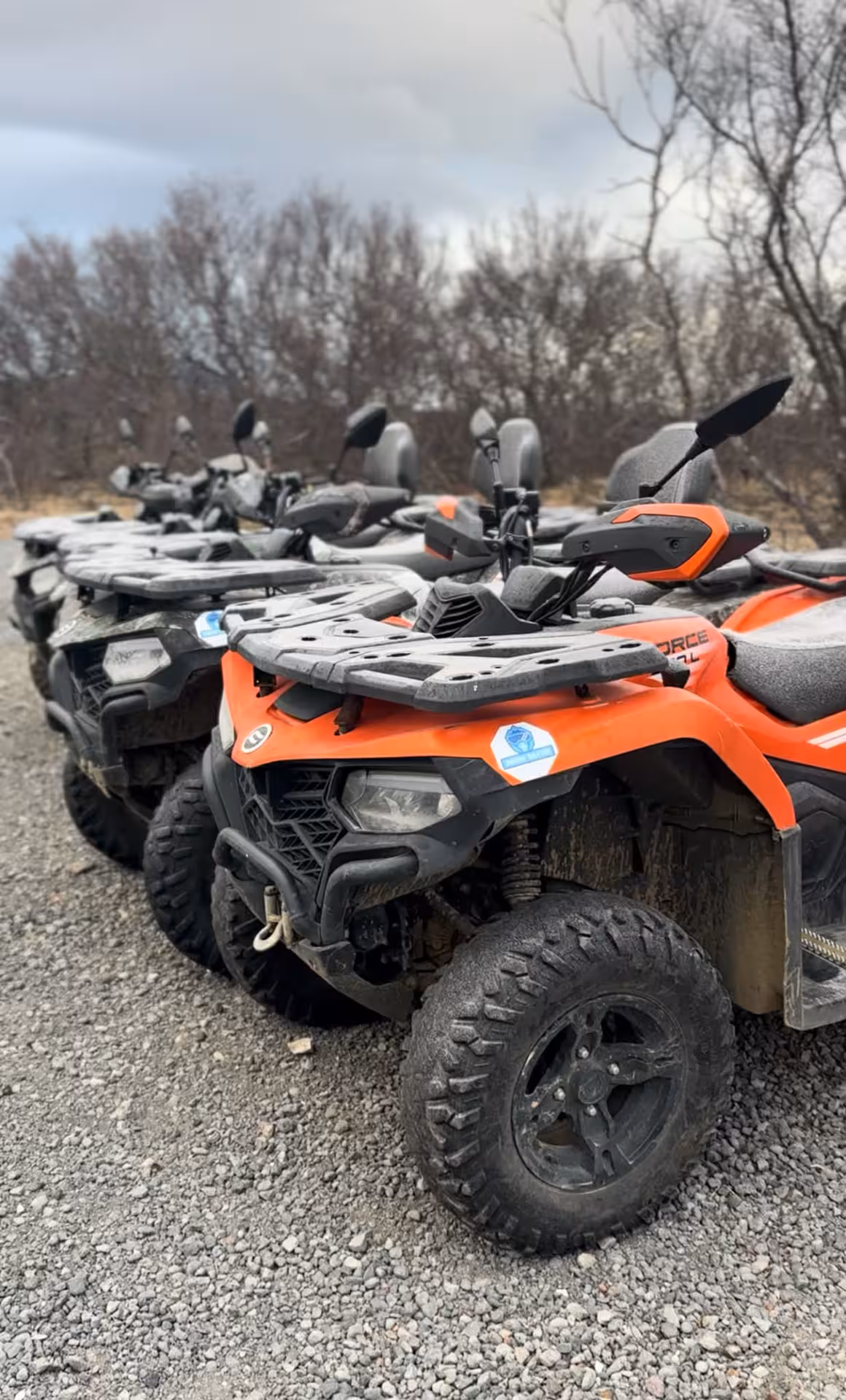 Row of orange CFMOTO quad bikes parked on gravel with bare trees in background