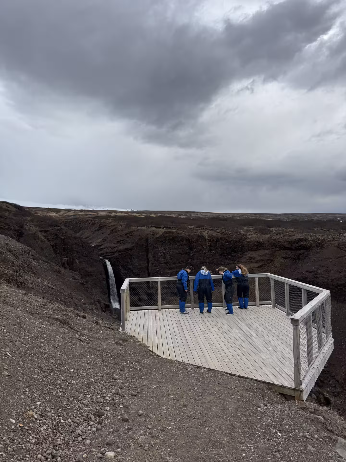 Four visitors in blue suits on a wooden viewing platform overlooking a waterfall and canyon in Husafell, Iceland