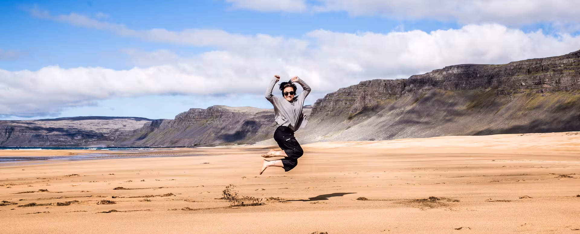 Person jumping joyfully on a sandy beach with dramatic cliffs and fjord in Iceland