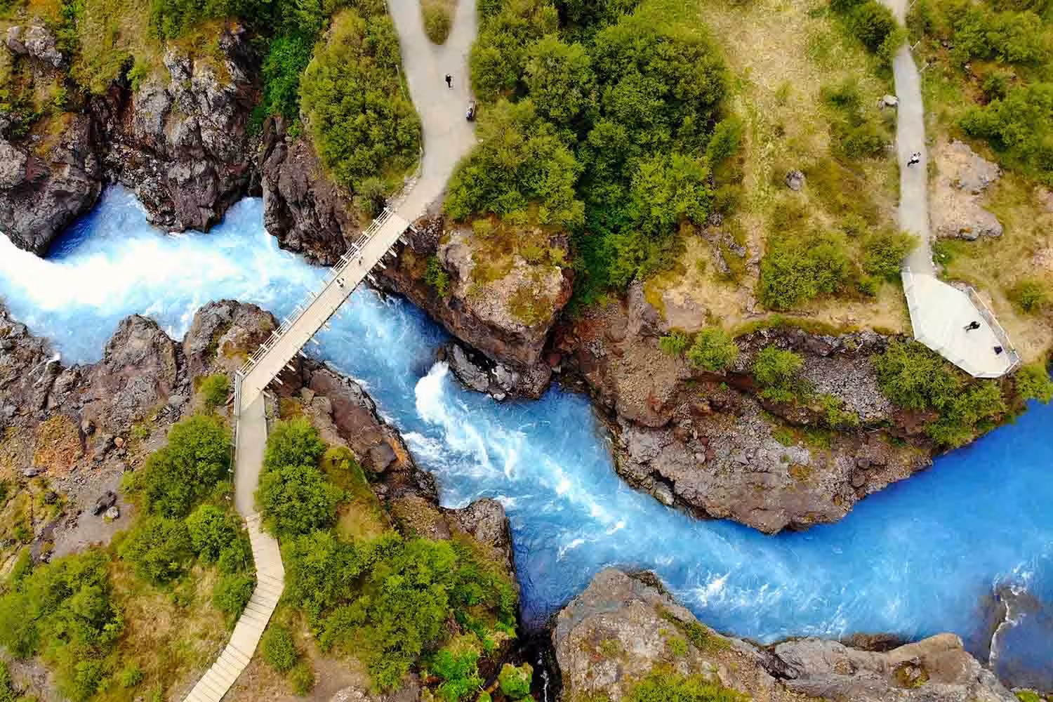 Aerial view of turquoise glacial river rushing through rocky canyon with footbridge in Husafell Iceland