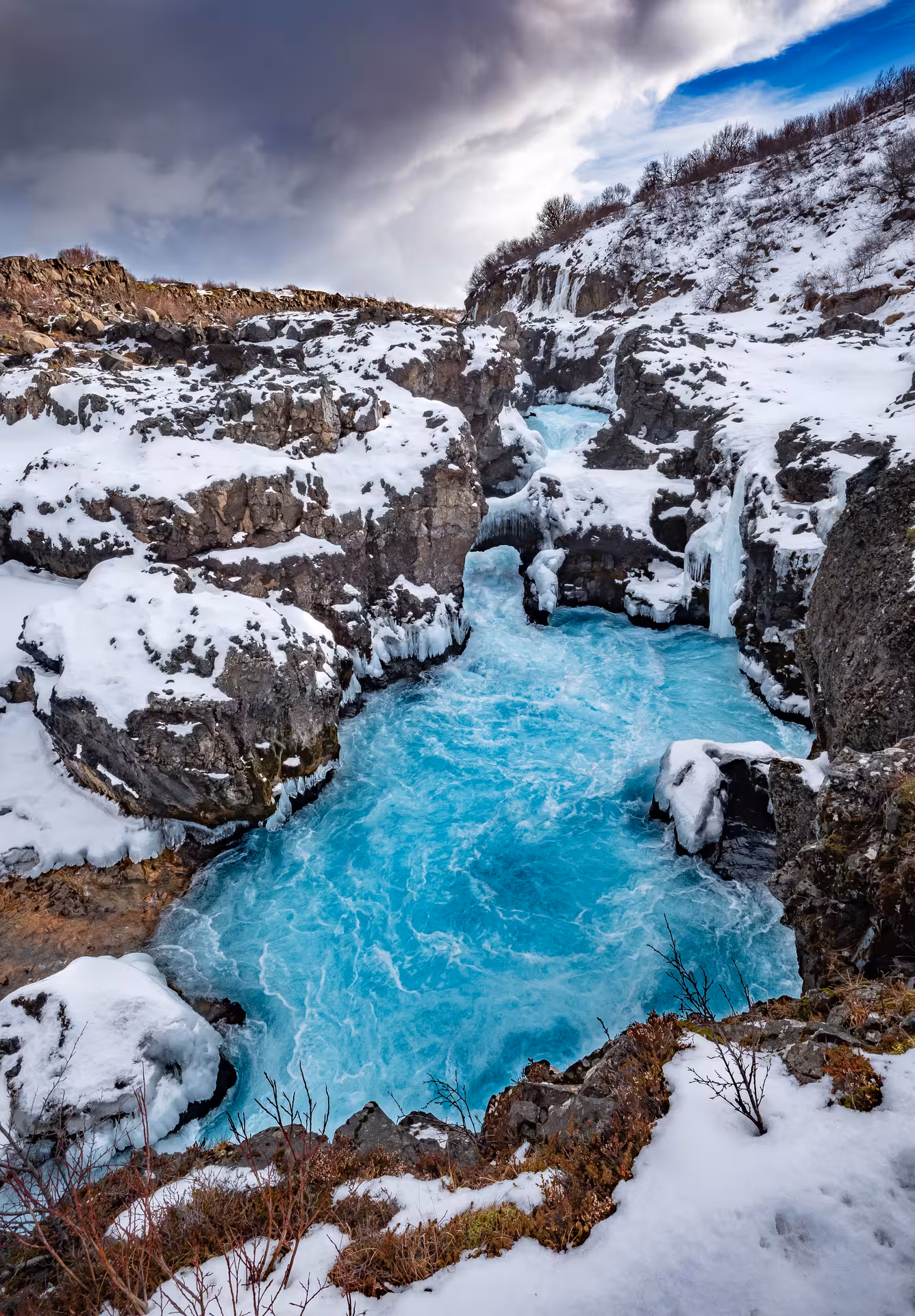 Vivid turquoise glacial river rushing through snow-covered lava rock canyon in Husafell, Iceland in winter