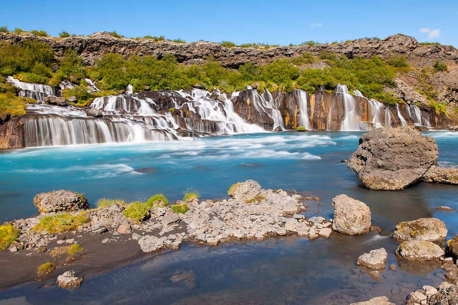 Multiple cascading waterfalls flowing into a turquoise glacial river with rocky lava formations in Iceland