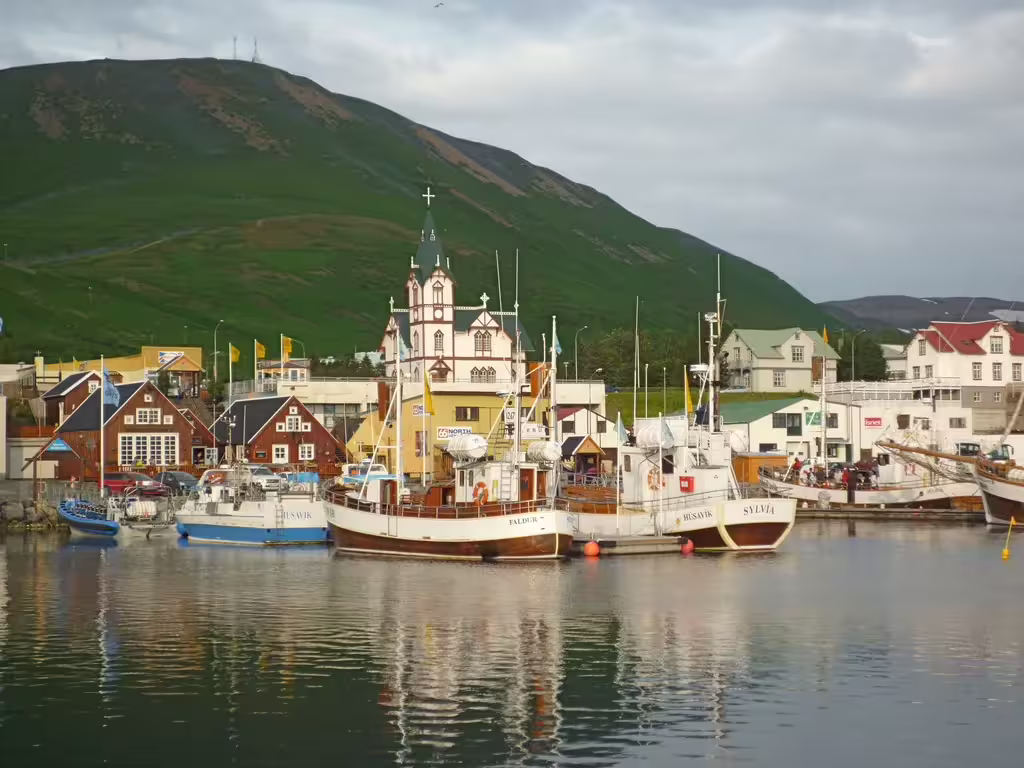 Húsavík harbor with fishing boats, colorful buildings, church steeple, and green mountain backdrop in Iceland