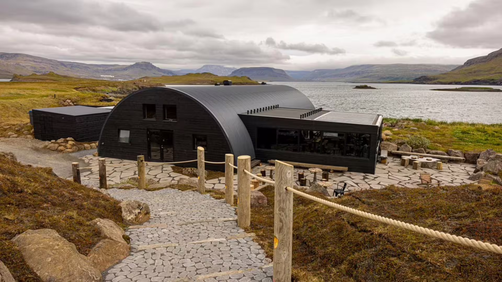 Black curved arch building at Hvammsvik Hot Springs beside a calm fjord with mountains in background