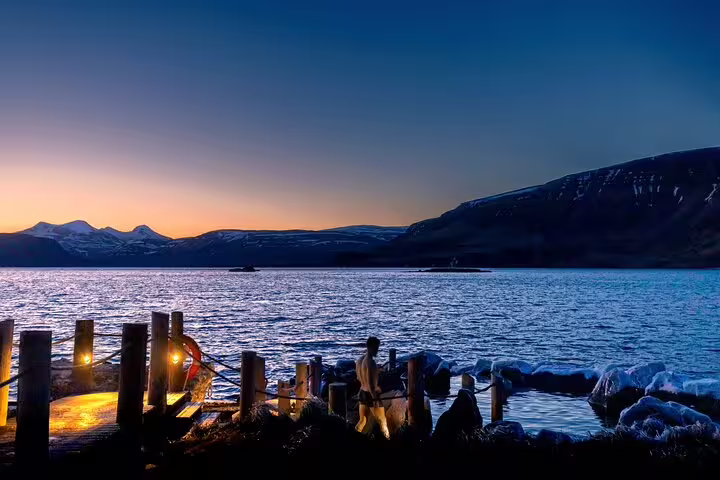 Hvammsvik Hot Springs at dusk with glowing lanterns on dock, fjord and snow-capped mountains in background