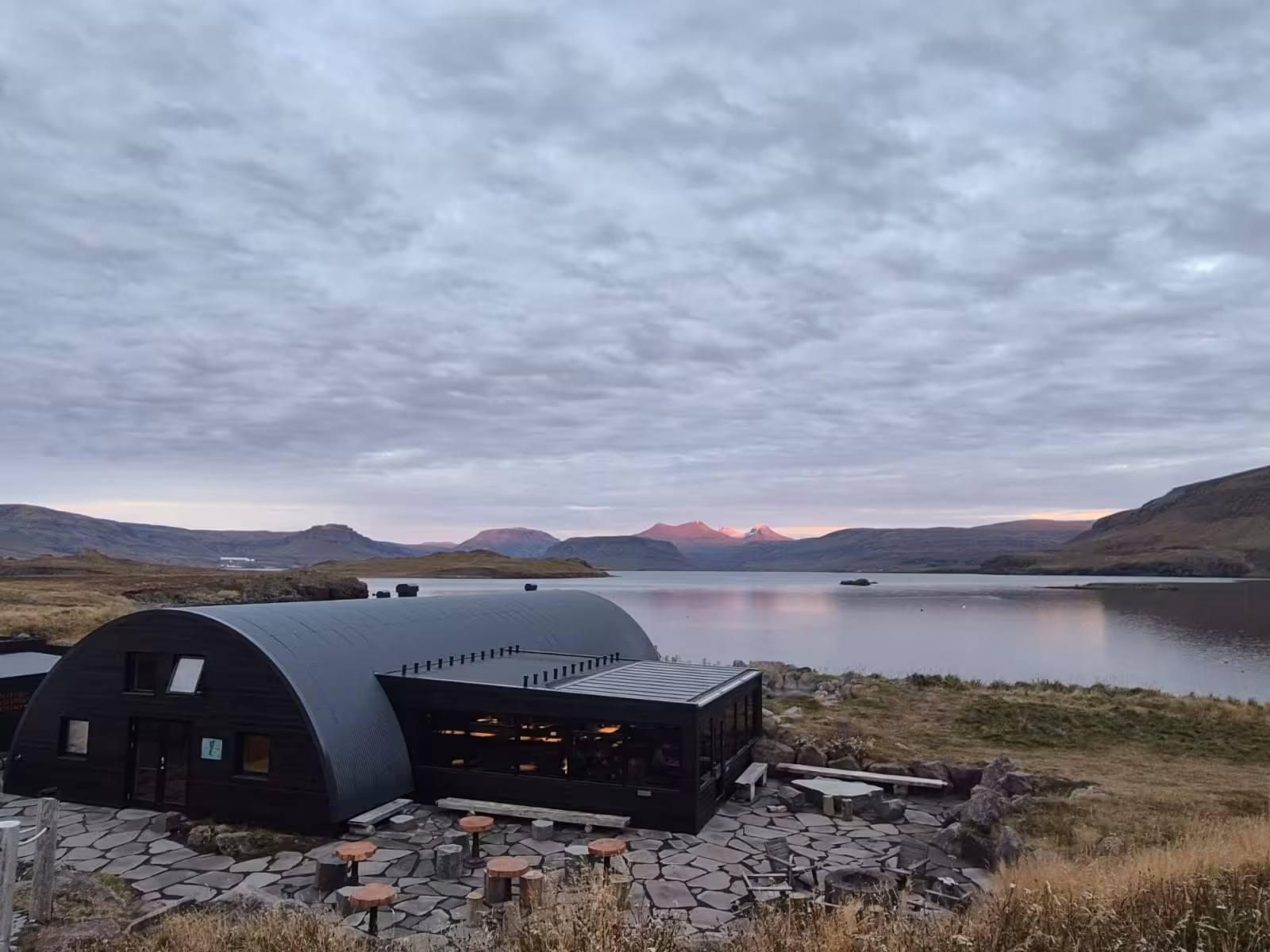 Dark curved building beside a calm lake at dusk with pink-lit mountains in Iceland