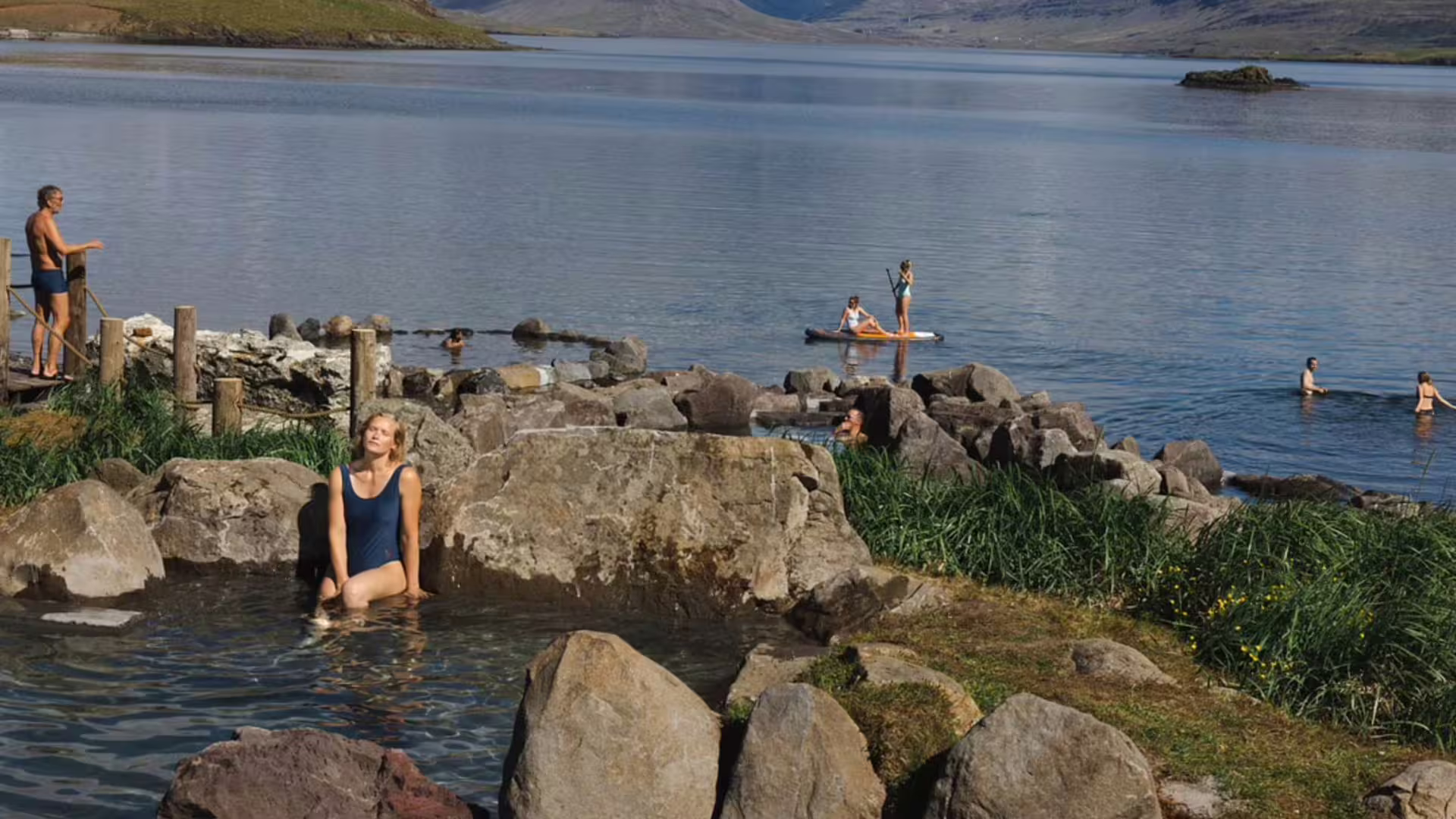 Visitors relaxing in natural hot spring pools at Hvammsvik beside a calm fjord with paddleboarders in Iceland