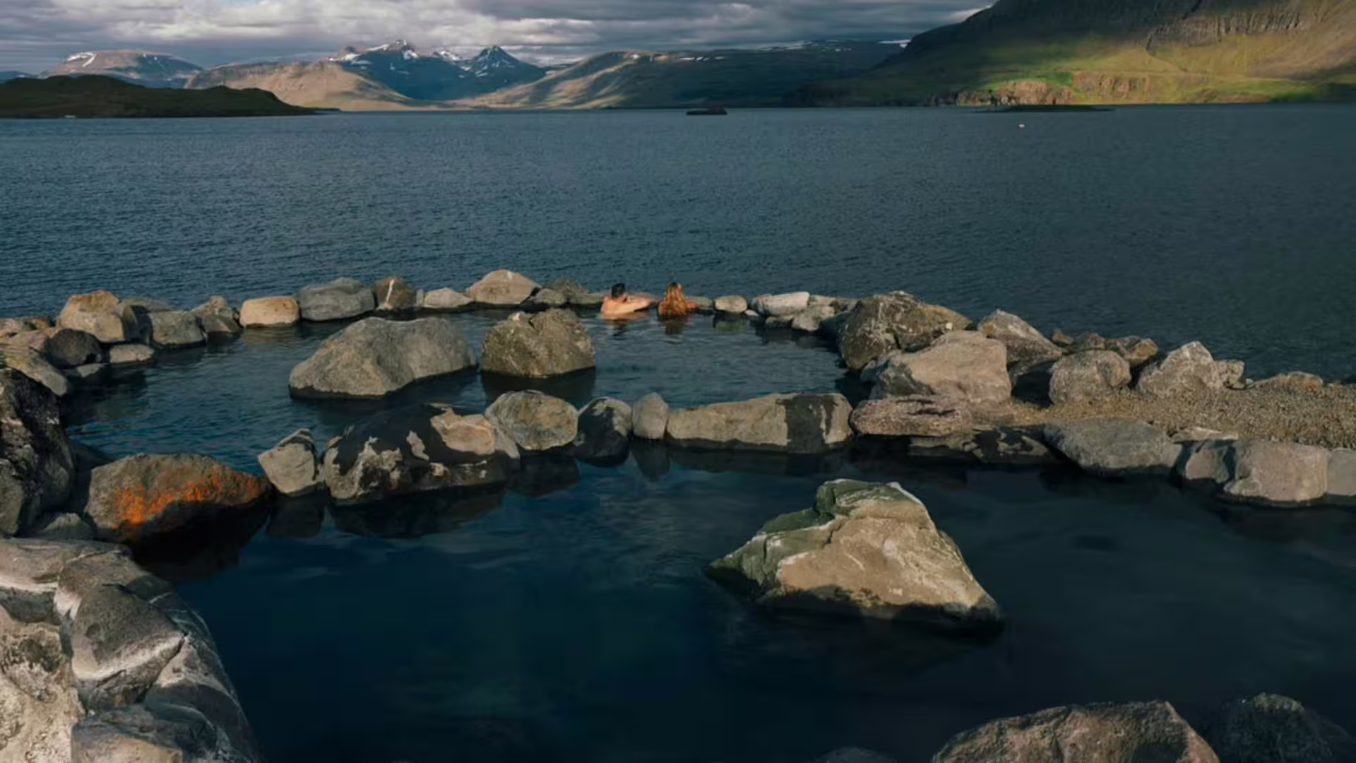 Bathers relaxing in a natural hot spring pool made of rocks beside a fjord with snow-capped mountains in Iceland
