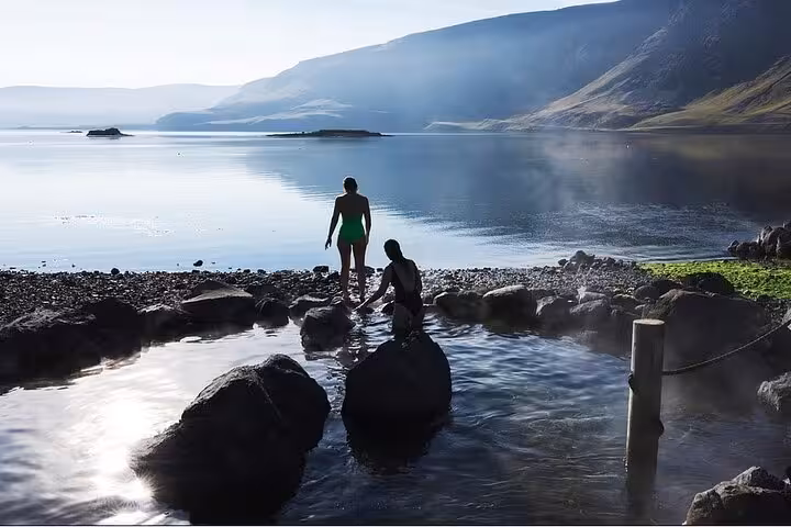 Two visitors enjoying Hvammsvík Hot Springs by a calm fjord with misty mountains in Iceland