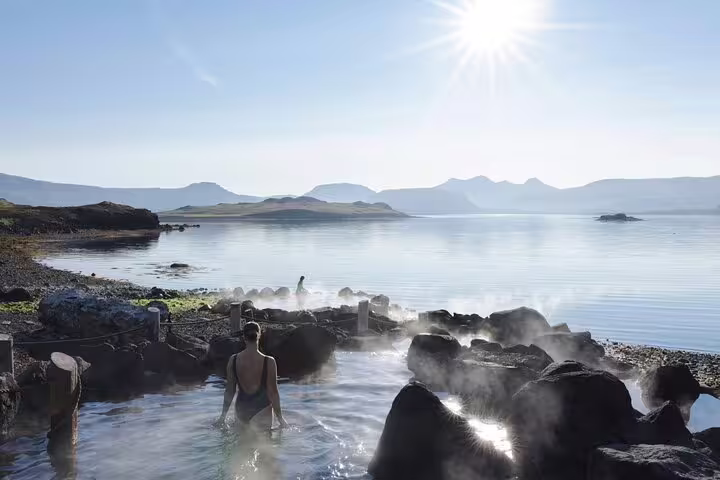 Steaming geothermal hot springs at Hvammsvik with rocky shores, calm fjord waters, and mountains in background
