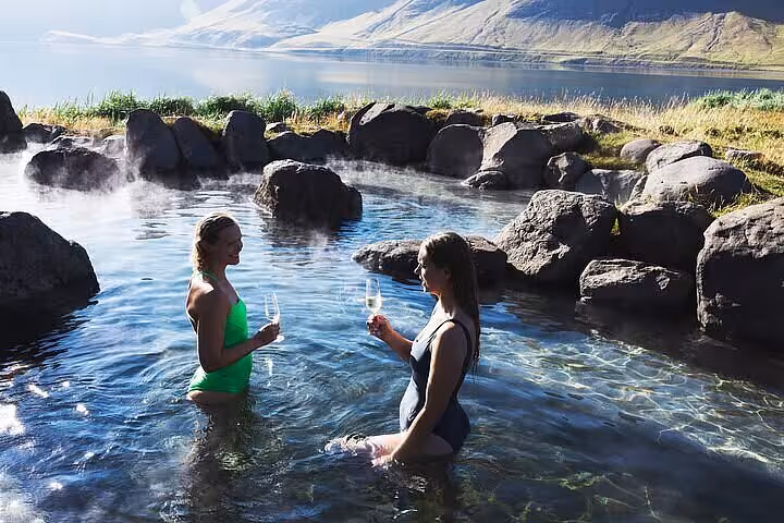 Two women in swimsuits toasting with champagne in Hvammsvík hot springs with scenic Icelandic fjord backdrop