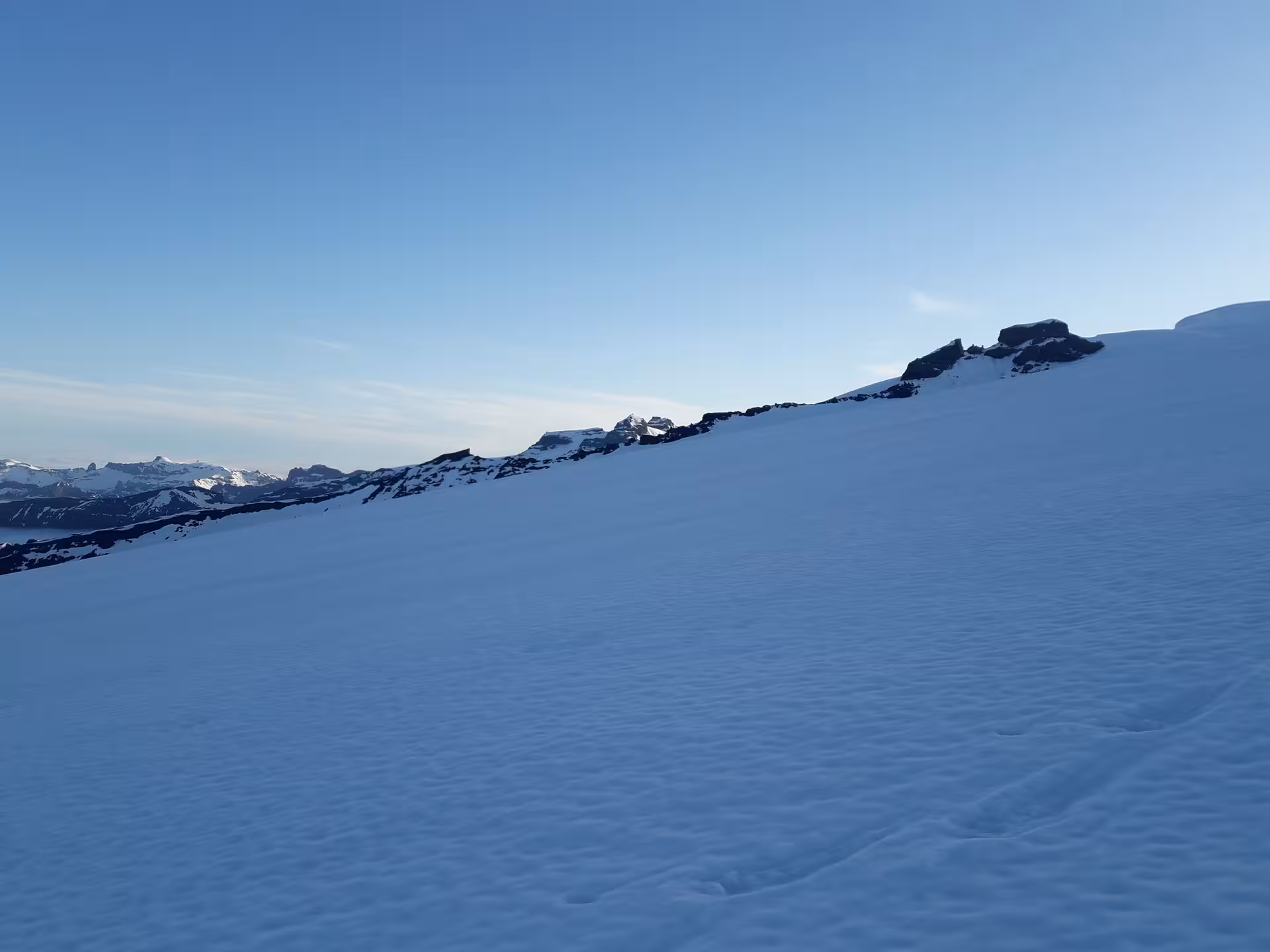 Snow-covered glacier slope with rocky outcrops and mountain peaks under clear blue sky in Iceland