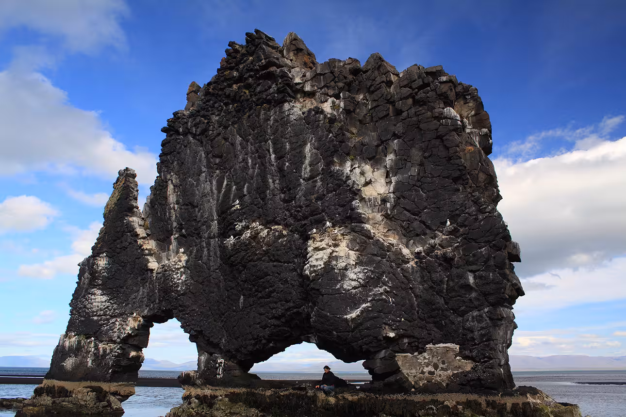 Massive black basalt rock formation Hvítserkur rising from the sea in North Iceland with a person sitting beneath it for scale