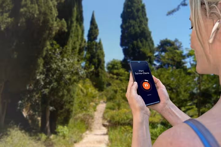 Woman holding smartphone with Parc Sainte-Claire audio tour app on a sunny garden path in Hyères