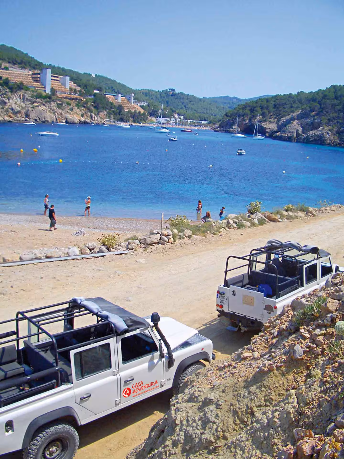 4x4 jeeps on an Ibiza Jeep Safari by a turquoise bay with boats, sandy track and coastal cliffs