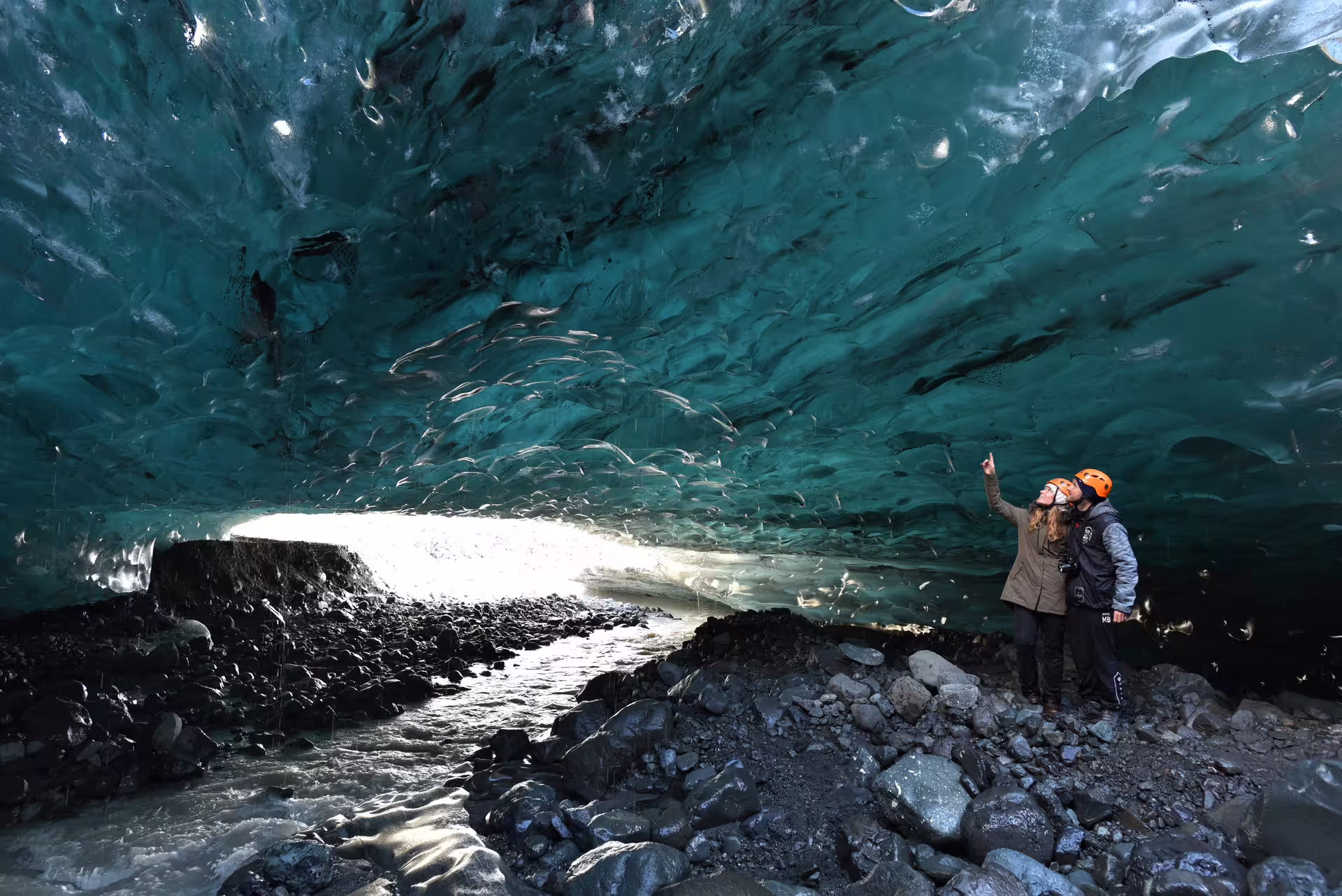Two tourists in orange helmets exploring a stunning blue ice cave with rocky floor in Iceland