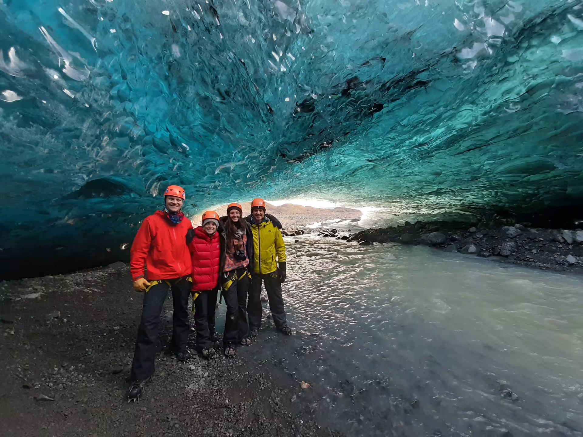 Four tourists in orange helmets posing inside a stunning blue glacial ice cave with a meltwater stream