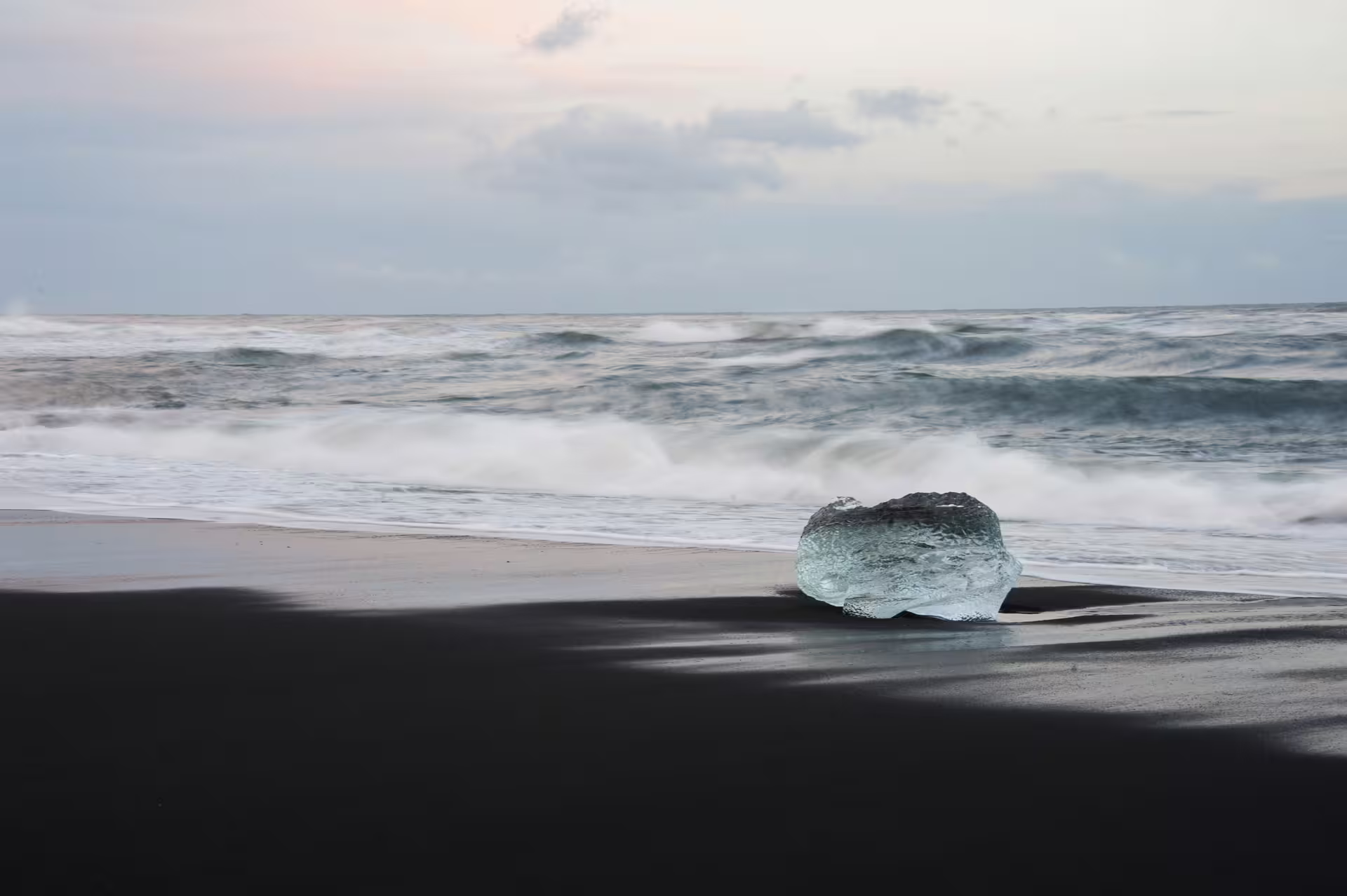 A translucent ice chunk resting on Iceland's black sand beach with waves crashing in the background