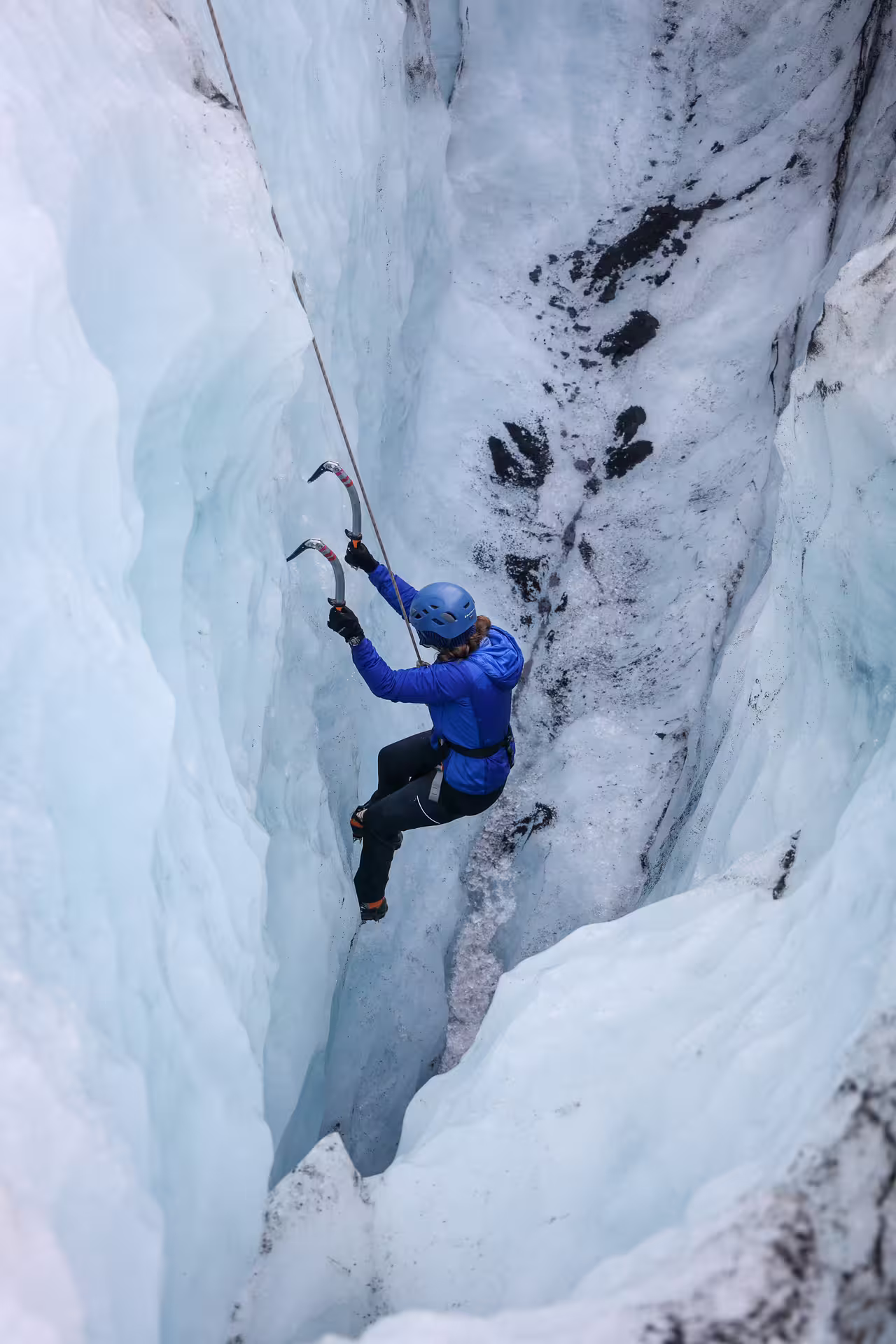 A climber in a blue helmet and jacket ascends a narrow ice crevasse with ice axes on Solheimajokull Glacier