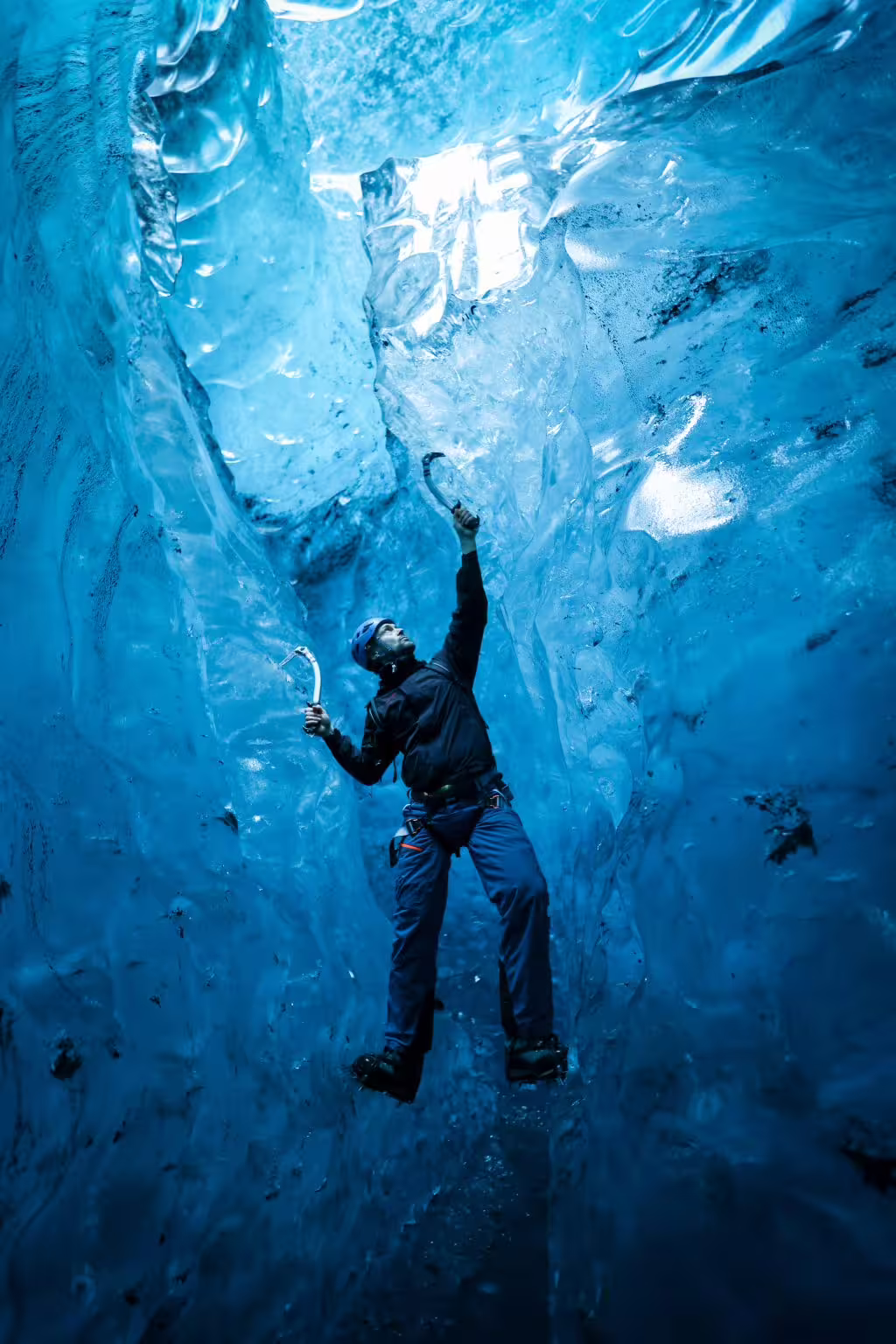 Ice climber with axes reaches upward inside a stunning blue glacial ice cave during a private climbing photoshoot adventure.