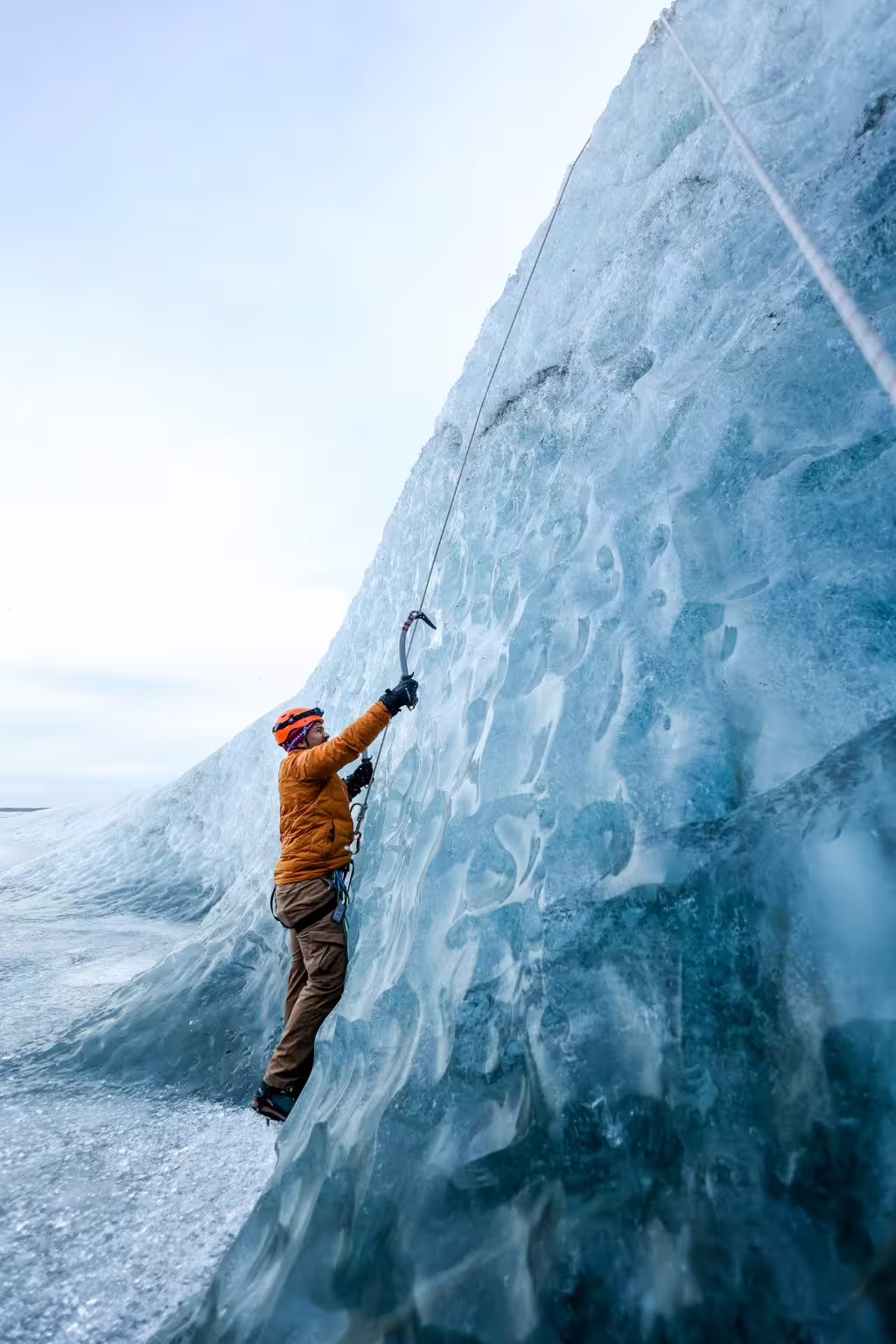 Ice climber in orange helmet scaling a towering blue glacial wall with ice axe on an adventure photoshoot