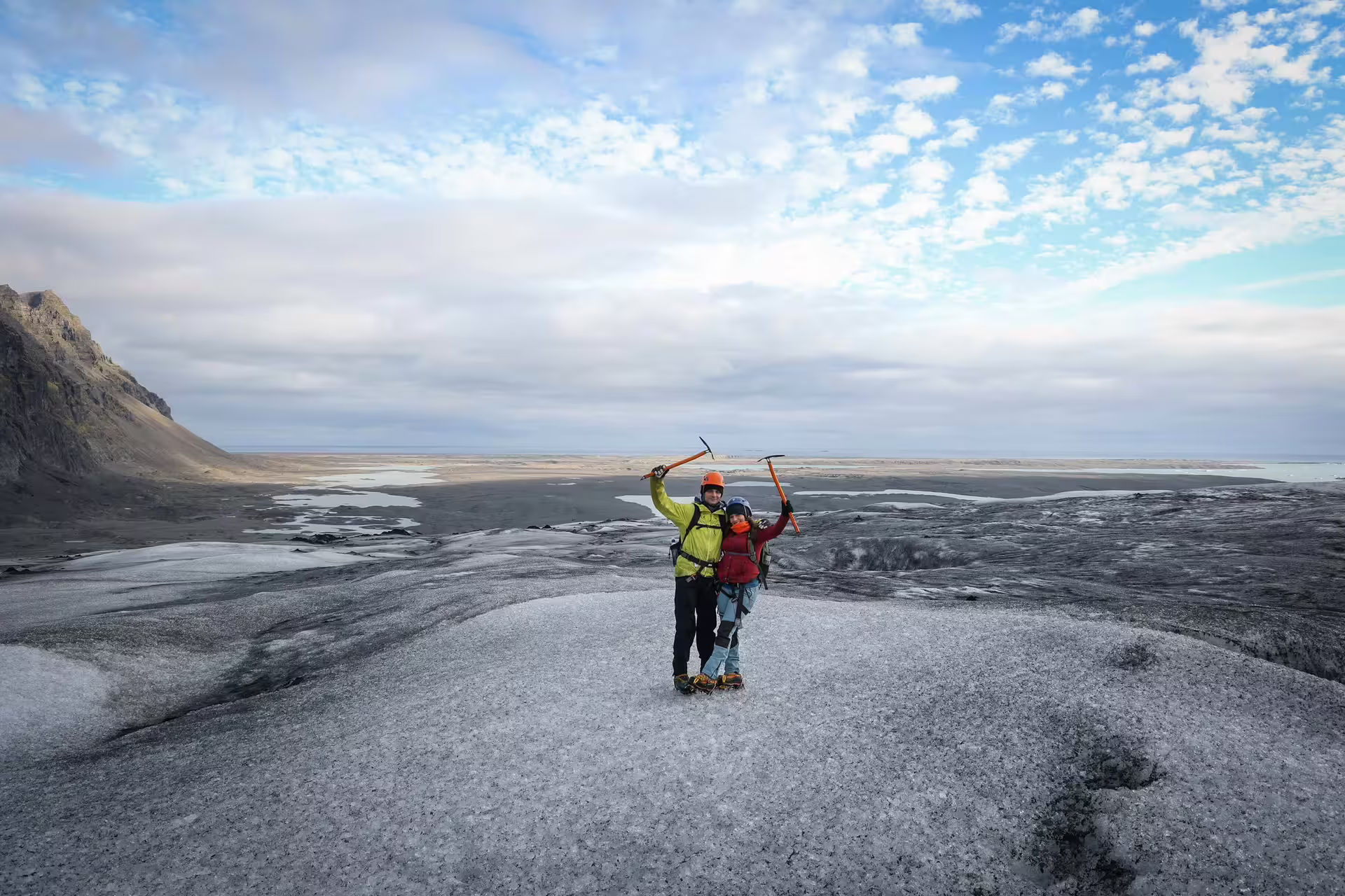 Two ice climbers celebrate with raised ice axes atop Skaftafell Glacier with Iceland's vast black sand plains behind them