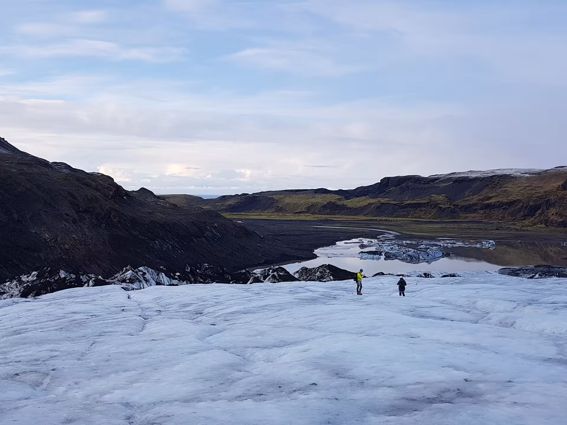 Two hikers on Sólheimajökull glacier in Iceland with glacial lagoon and black volcanic mountains behind