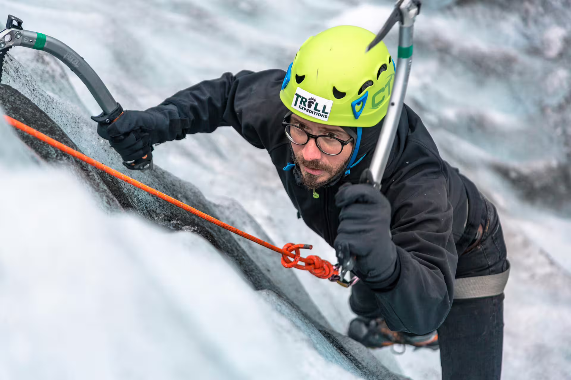 Ice climber scaling glacier wall with ice axes and orange rope at Skaftafell, wearing green Troll Expeditions helmet