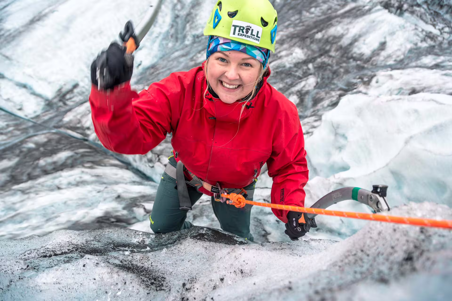 Smiling woman ice climbing on glacier face with ice axe and orange rope, wearing red jacket and yellow helmet