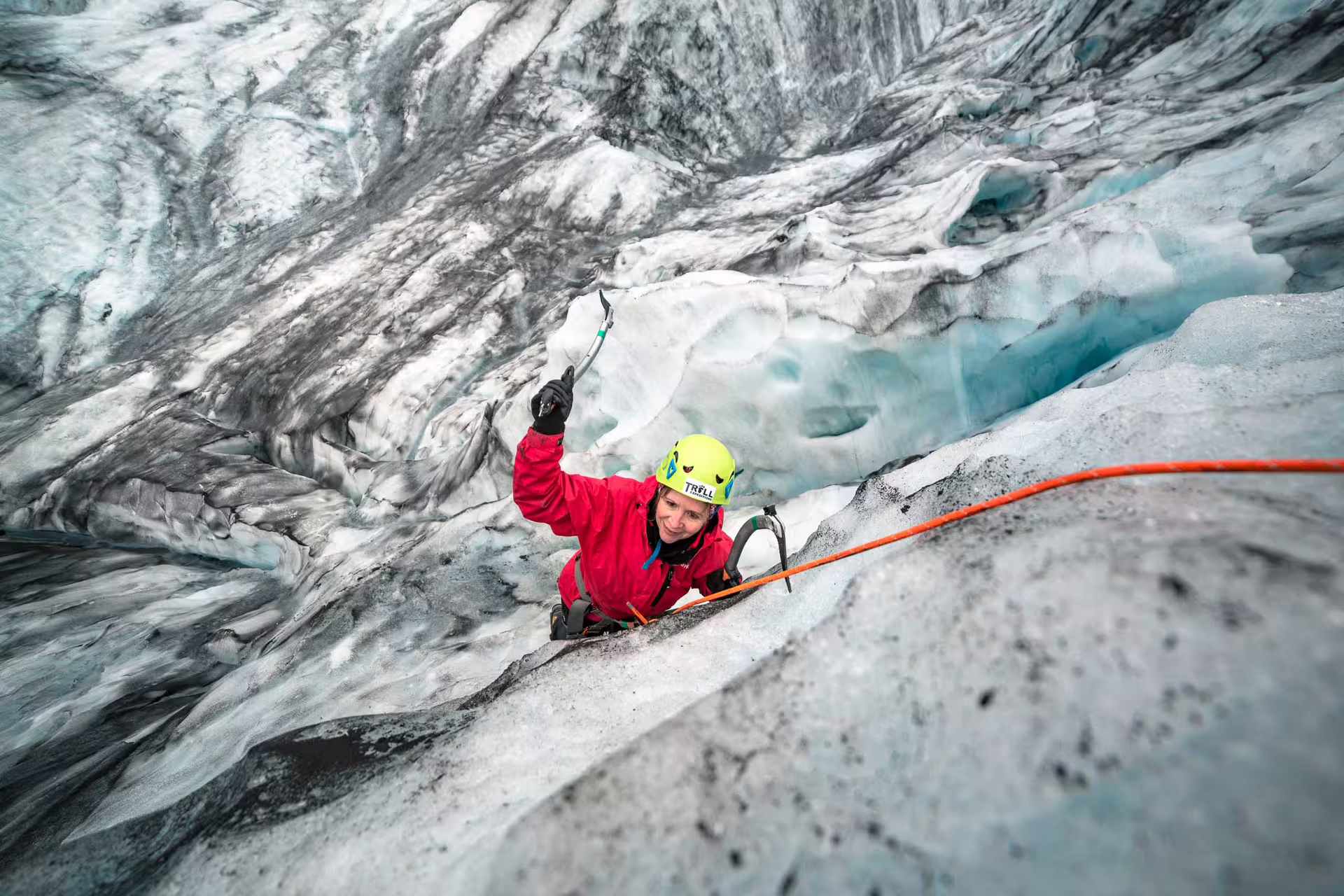 Smiling climber in red jacket ascending glacial ice wall with ice axes and orange rope at Skaftafell