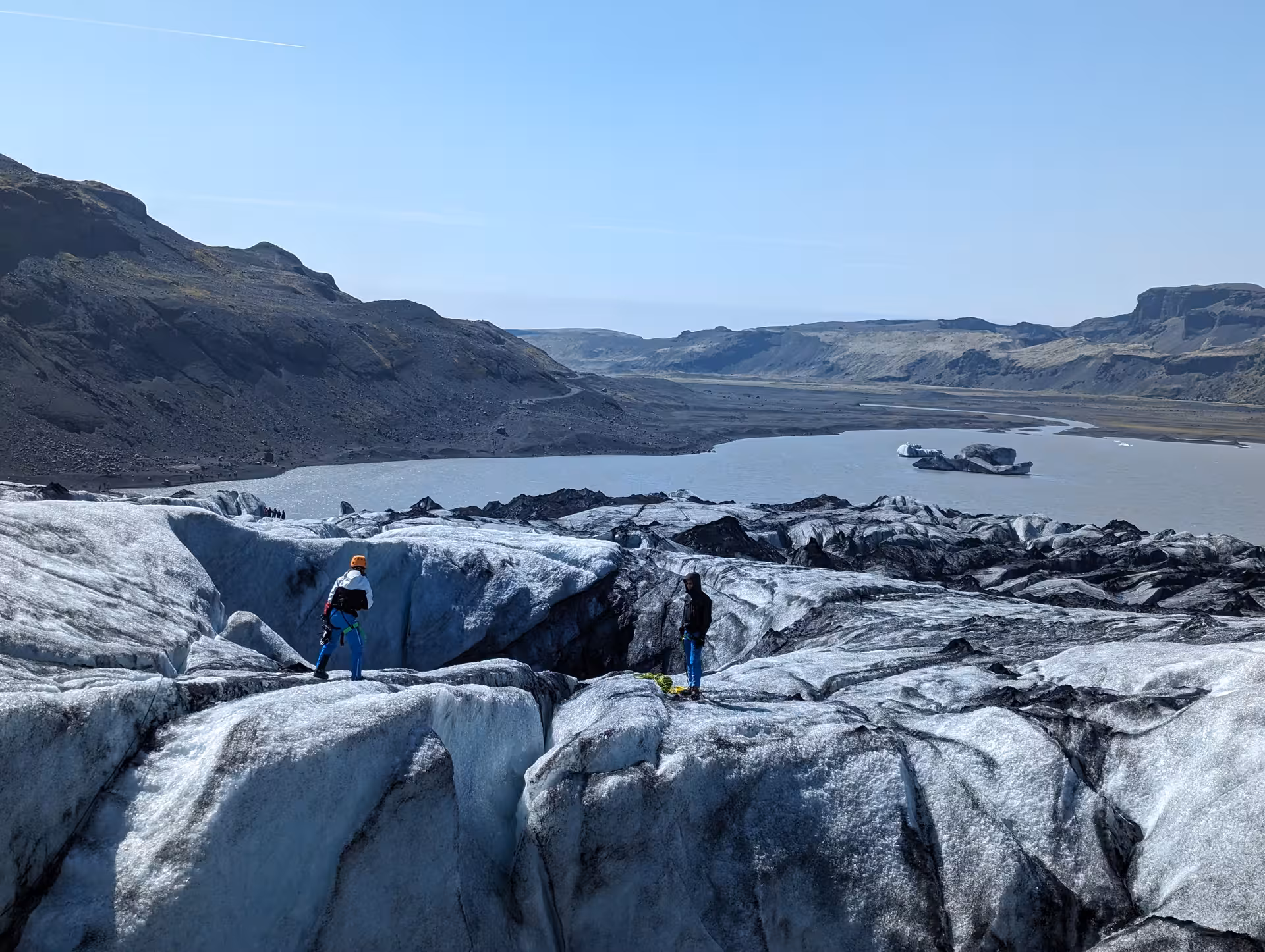 Two climbers with gear standing on crevassed Solheimajokull glacier with glacial lagoon and mountains behind