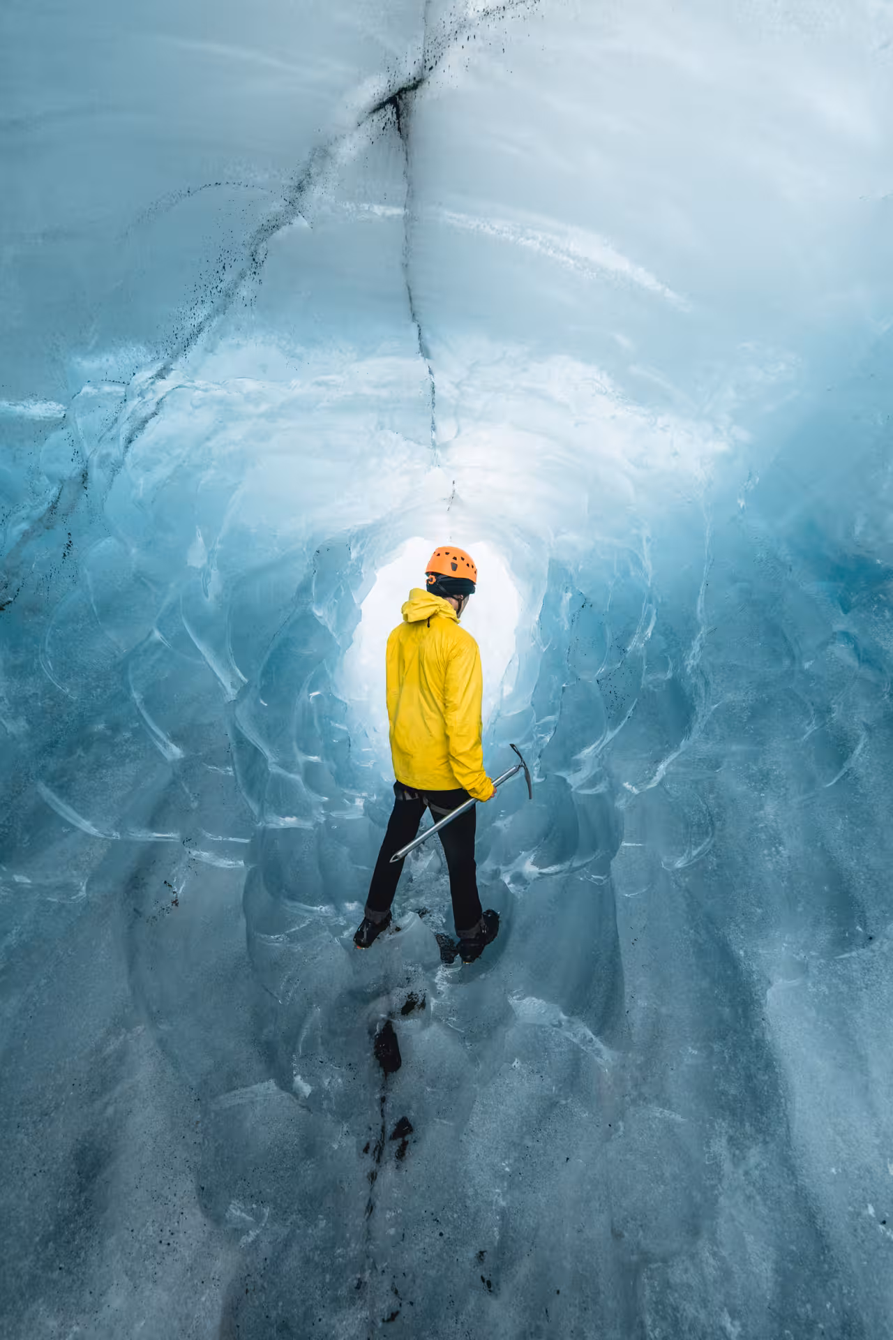 Explorer in yellow jacket and orange helmet holding ice axe inside blue glacial ice cave tunnel