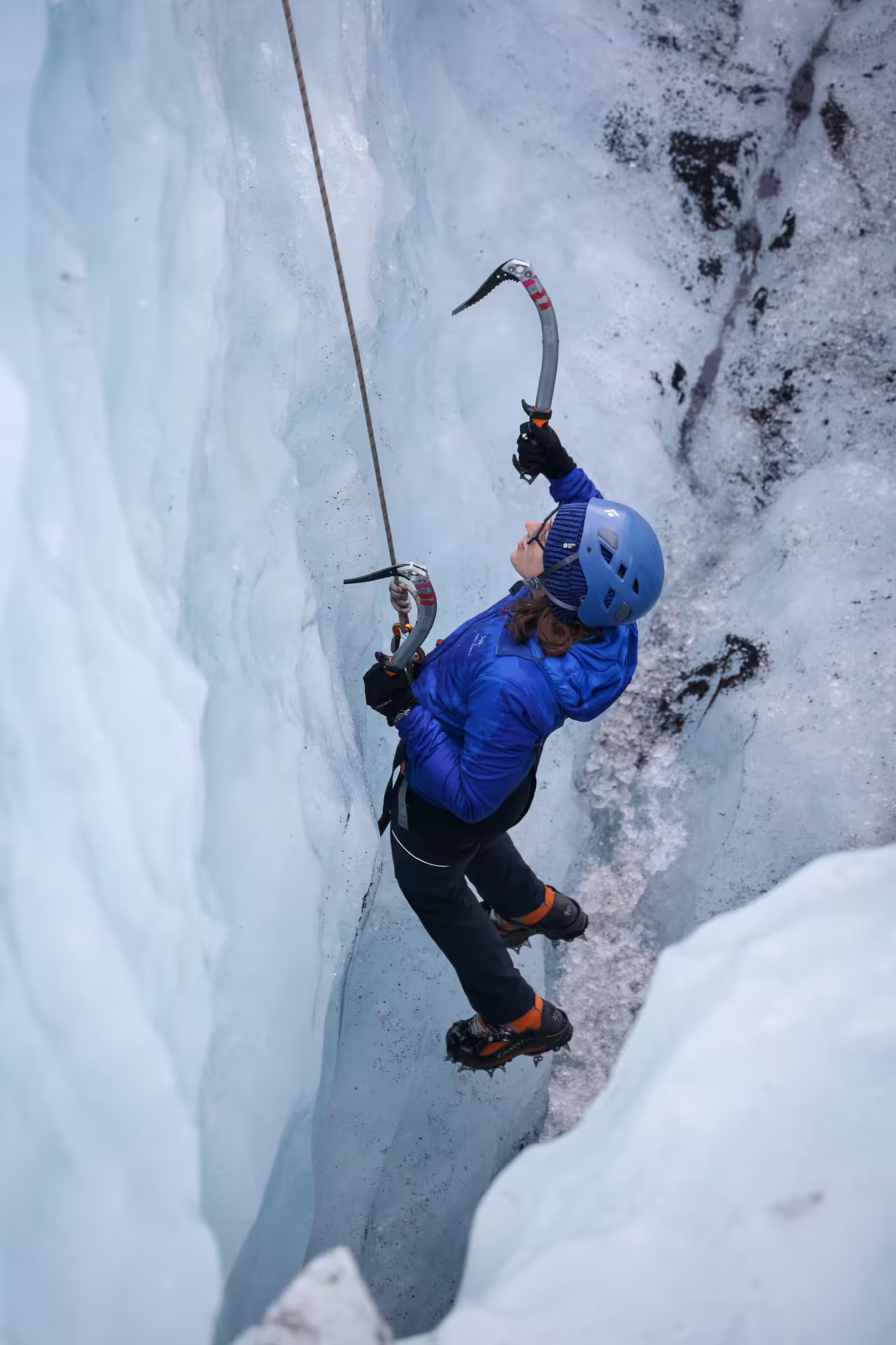 A climber in a blue jacket scales a steep ice wall with axes and crampons on Solheimajokull Glacier Iceland.