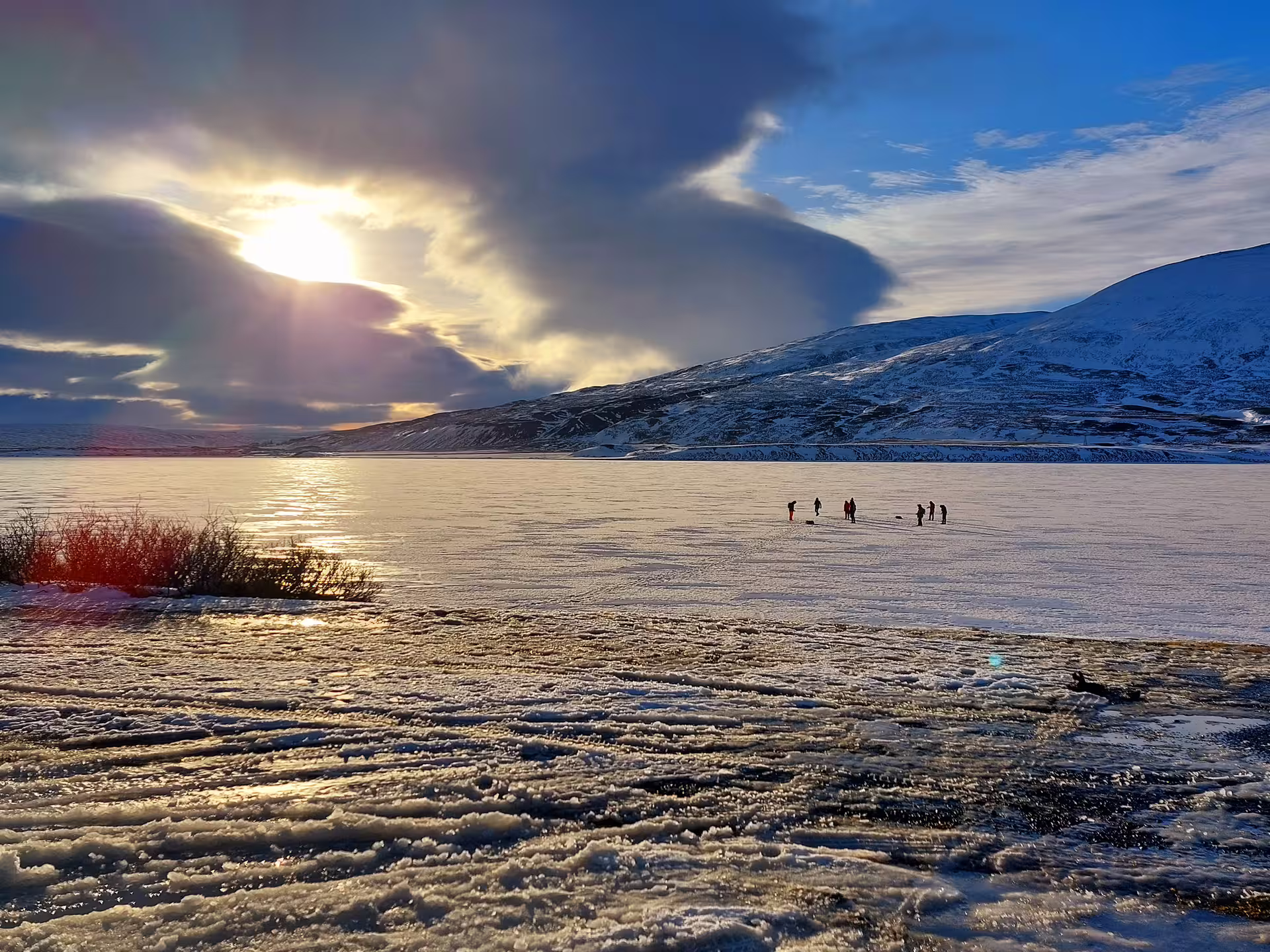 Group of people ice fishing on a frozen lake in Akureyri with snow-covered mountains at sunset
