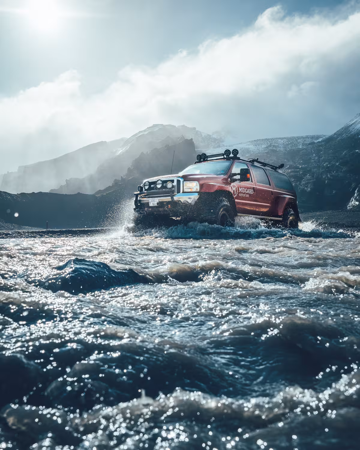Red Midgard Adventure 4x4 SUV fording a glacial river with snow-capped volcanic mountains in Iceland