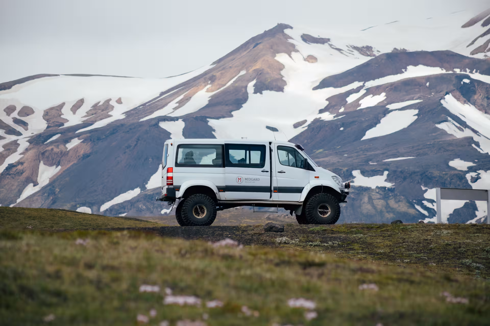 White Midgard 4x4 off-road van parked on green highland terrain with snow-capped volcanic mountain in Iceland