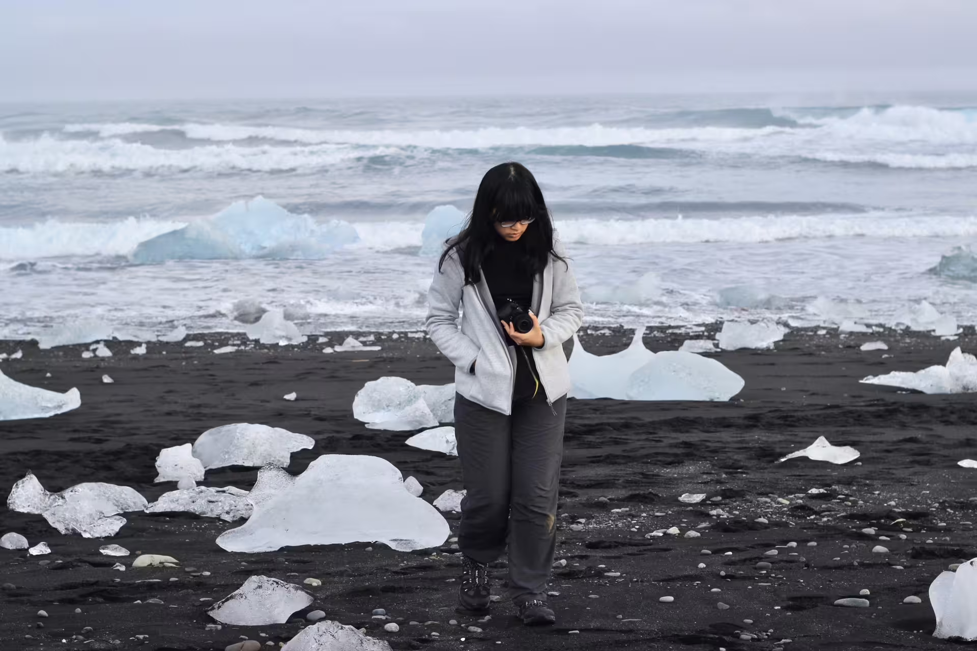 Photographer standing on Iceland's black sand beach surrounded by glacial ice chunks with ocean waves behind