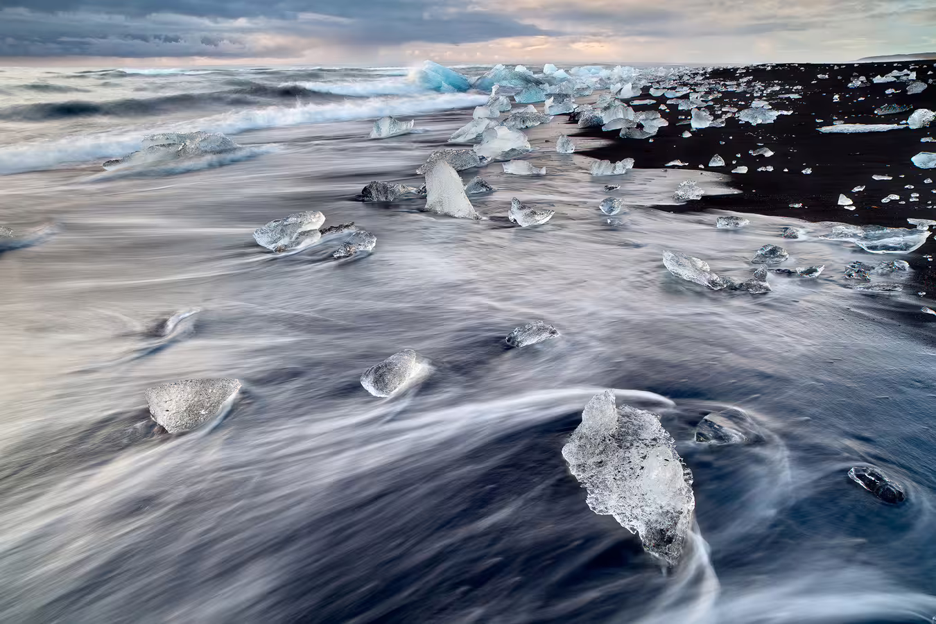 Ice chunks scattered on Iceland's black sand beach with waves washing ashore under dramatic cloudy skies