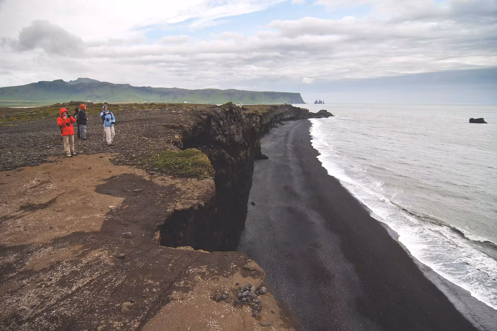 Tourists on black sand cliffs overlooking Iceland's volcanic south coast beach with ocean waves