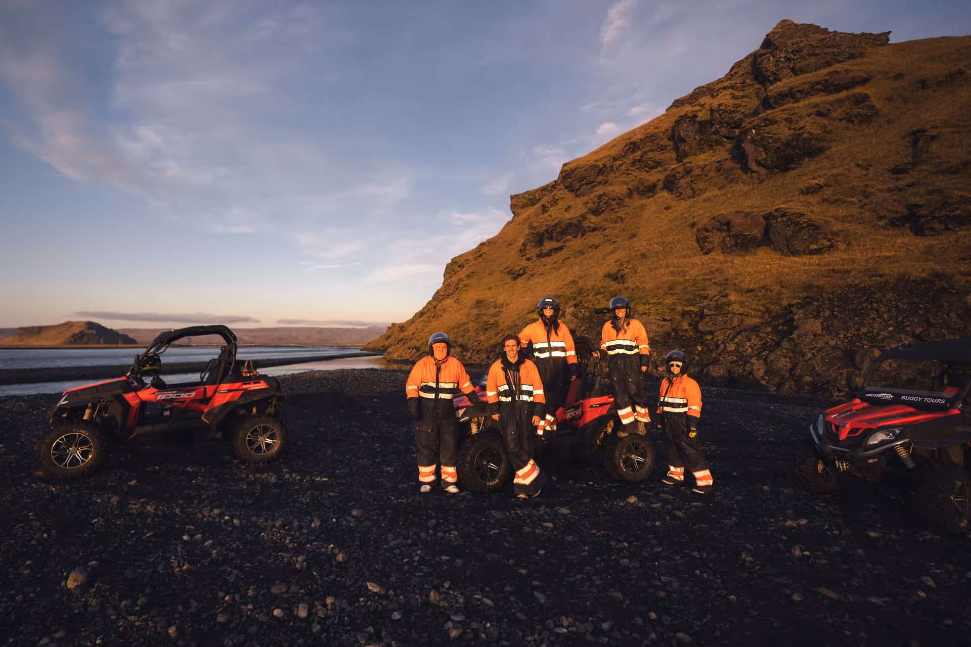 Group of five people in orange safety suits posing with red buggies on Iceland's black sand beach