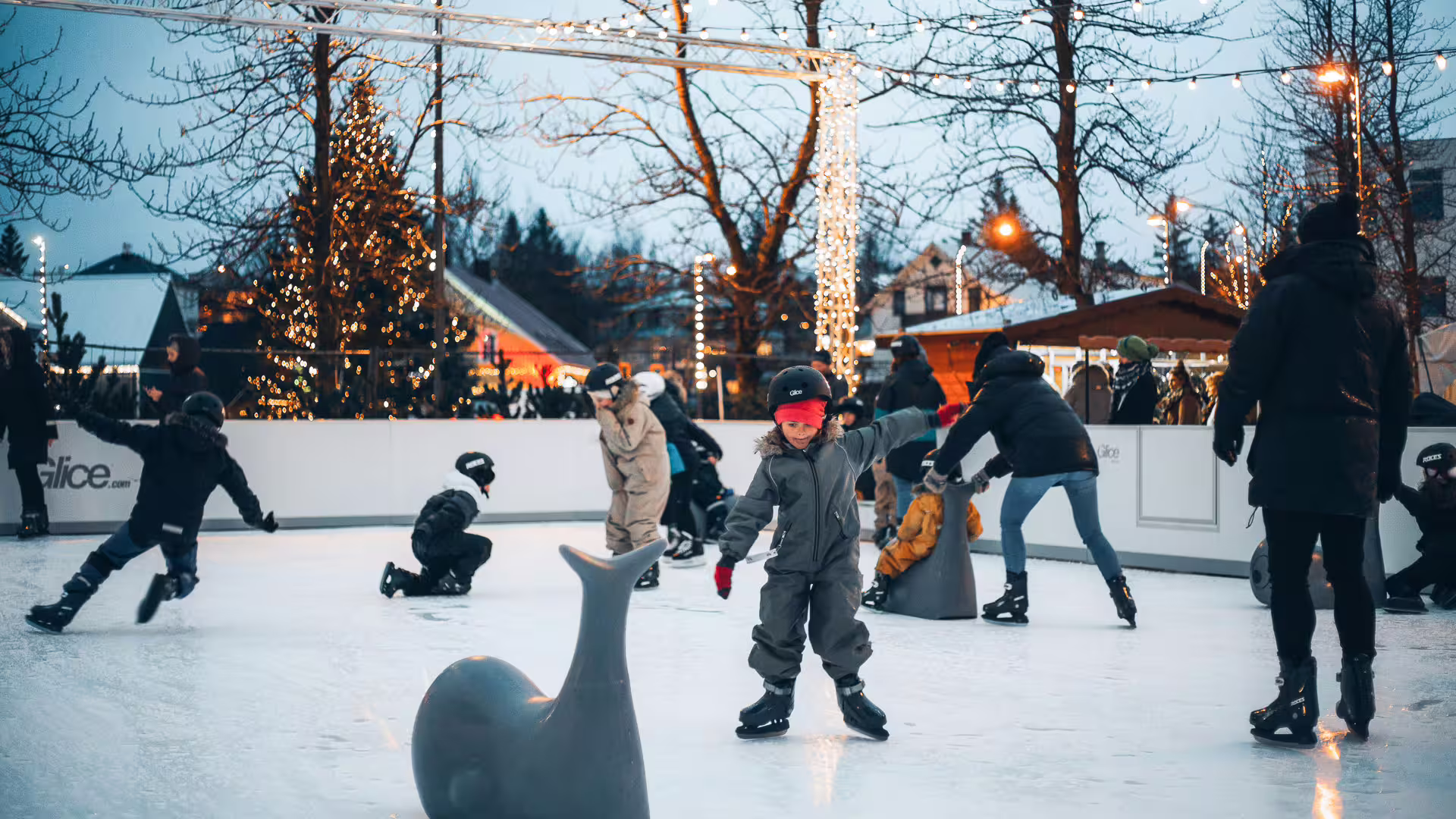 Children and adults ice skating on an outdoor rink with Christmas lights and festive decorations at dusk
