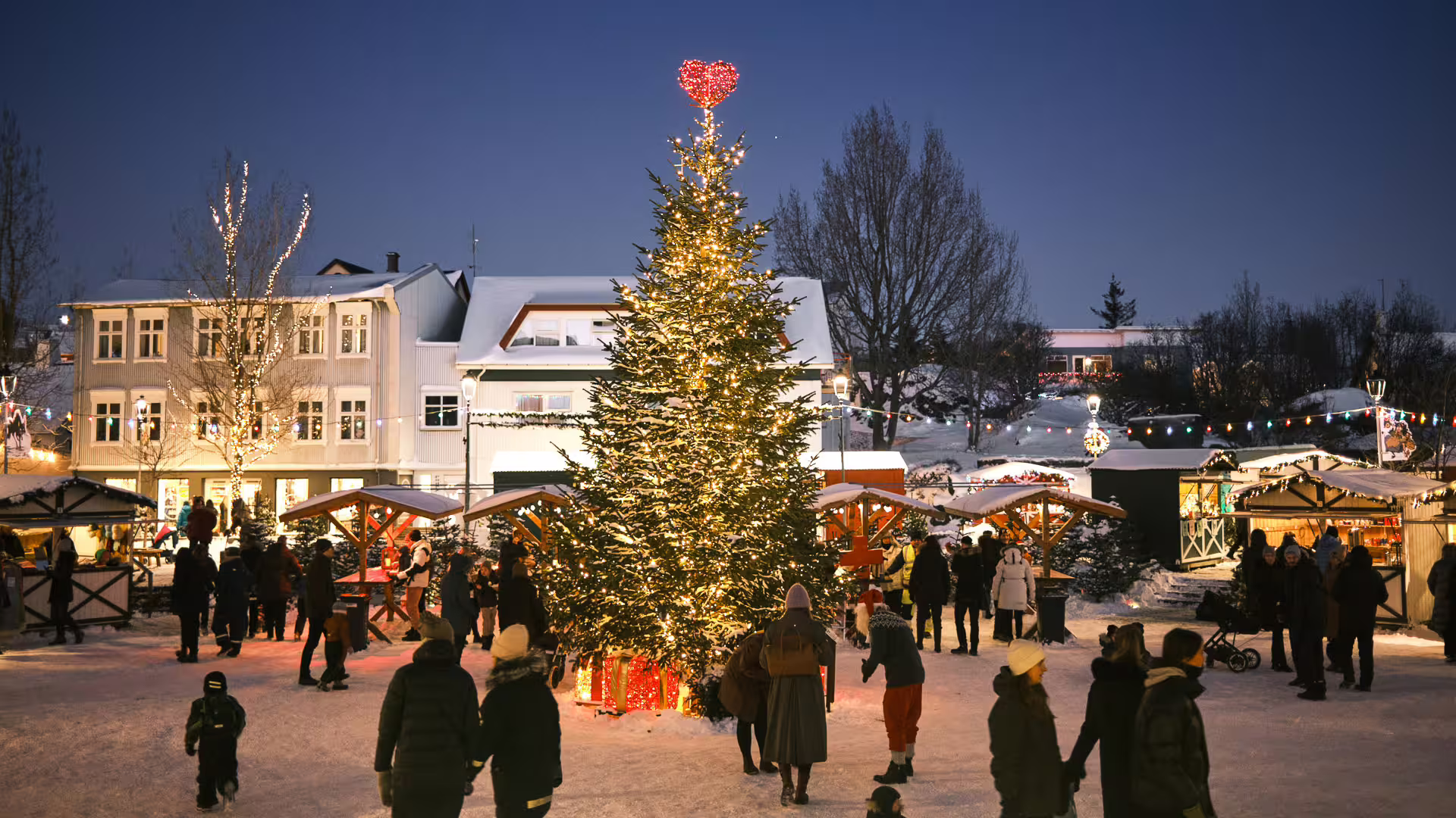 Festive Icelandic Christmas market at dusk with lit tree, heart topper, snowy stalls, and crowds of visitors