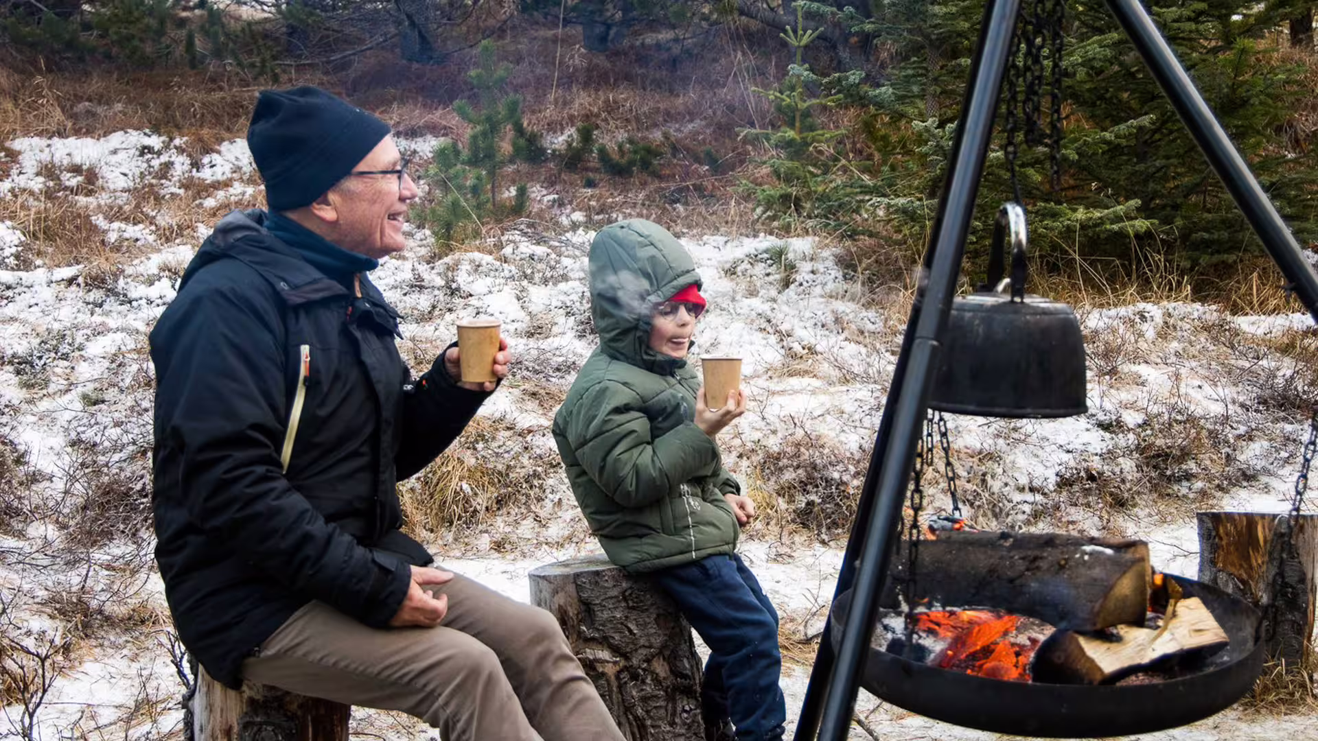 Man and child holding warm drinks by a campfire in a snowy Icelandic winter landscape
